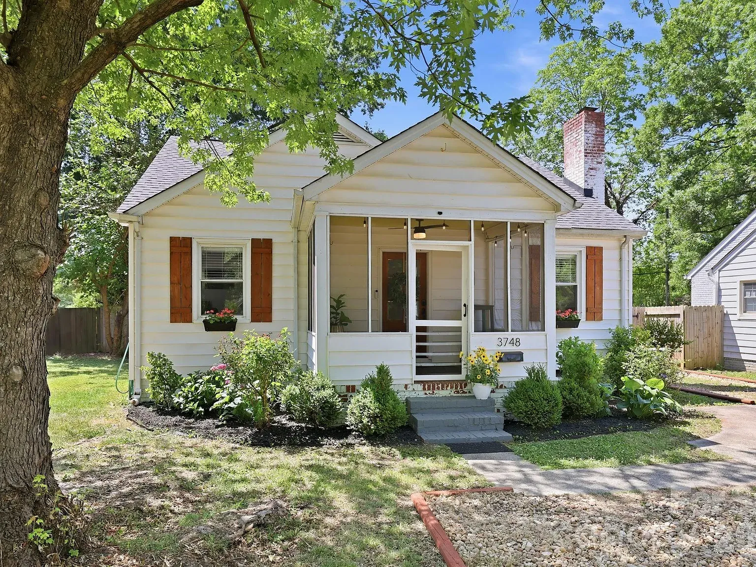 Small white house with a screened front porch, brown shutters, and a brick chimney. Front garden with flowers, a large tree in the foreground, and the number 3748 near the entry.