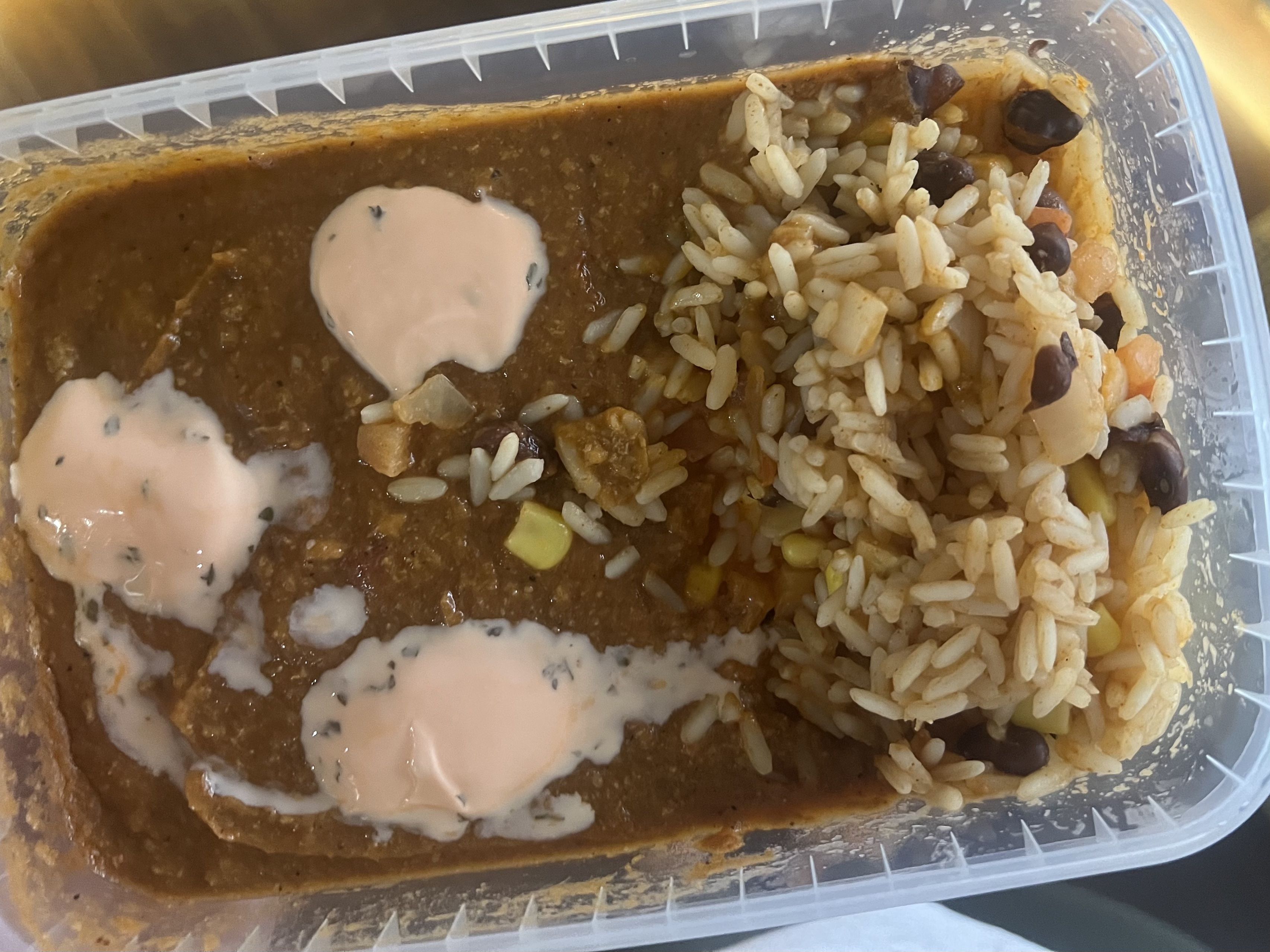 Plastic container with a turkey stew topped with three dollops of pale pink sauce, alongside white rice mixed with black beans, corn, and vegetables.