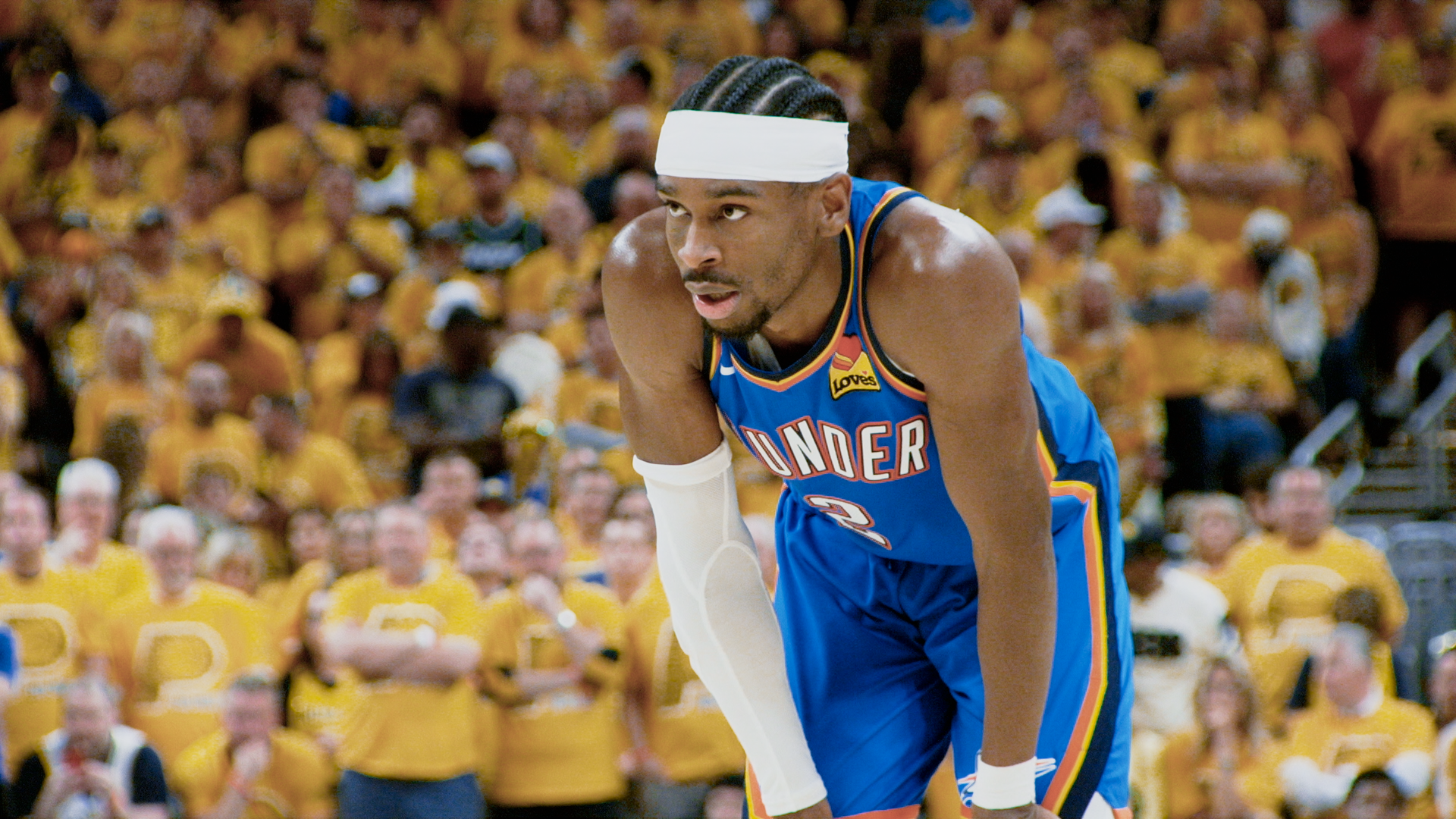Basketball player in blue Oklahoma City Thunder jersey, wearing white headband and arm sleeve, leaning forward on the court with a yellow-shirted crowd blurred in the background.
