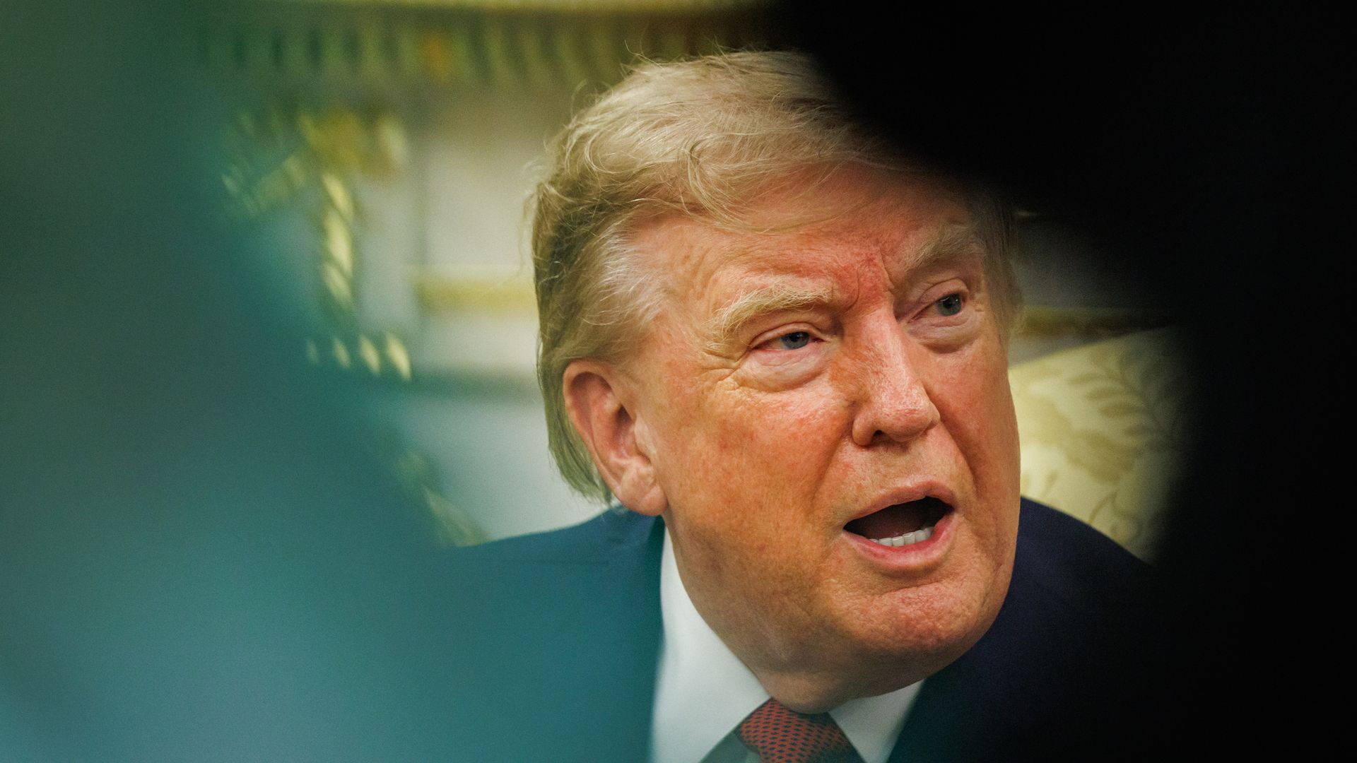Close-up of former President Donald Trump, mouth open, partially obscured by green blur on the left and dark background on the right.