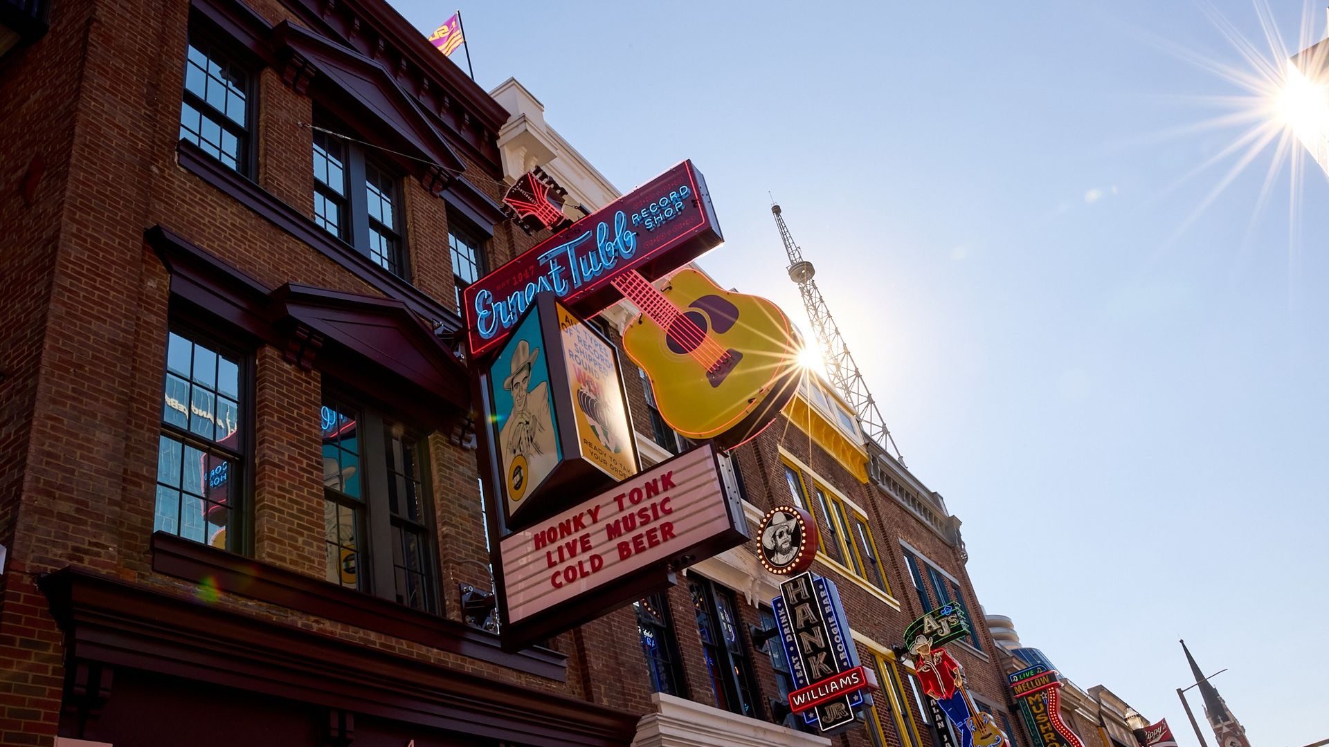 Sunlit street with brick buildings featuring colorful neon signs including a large yellow guitar and messages like "Honky Tonk Live Music Cold Beer" under clear blue sky.