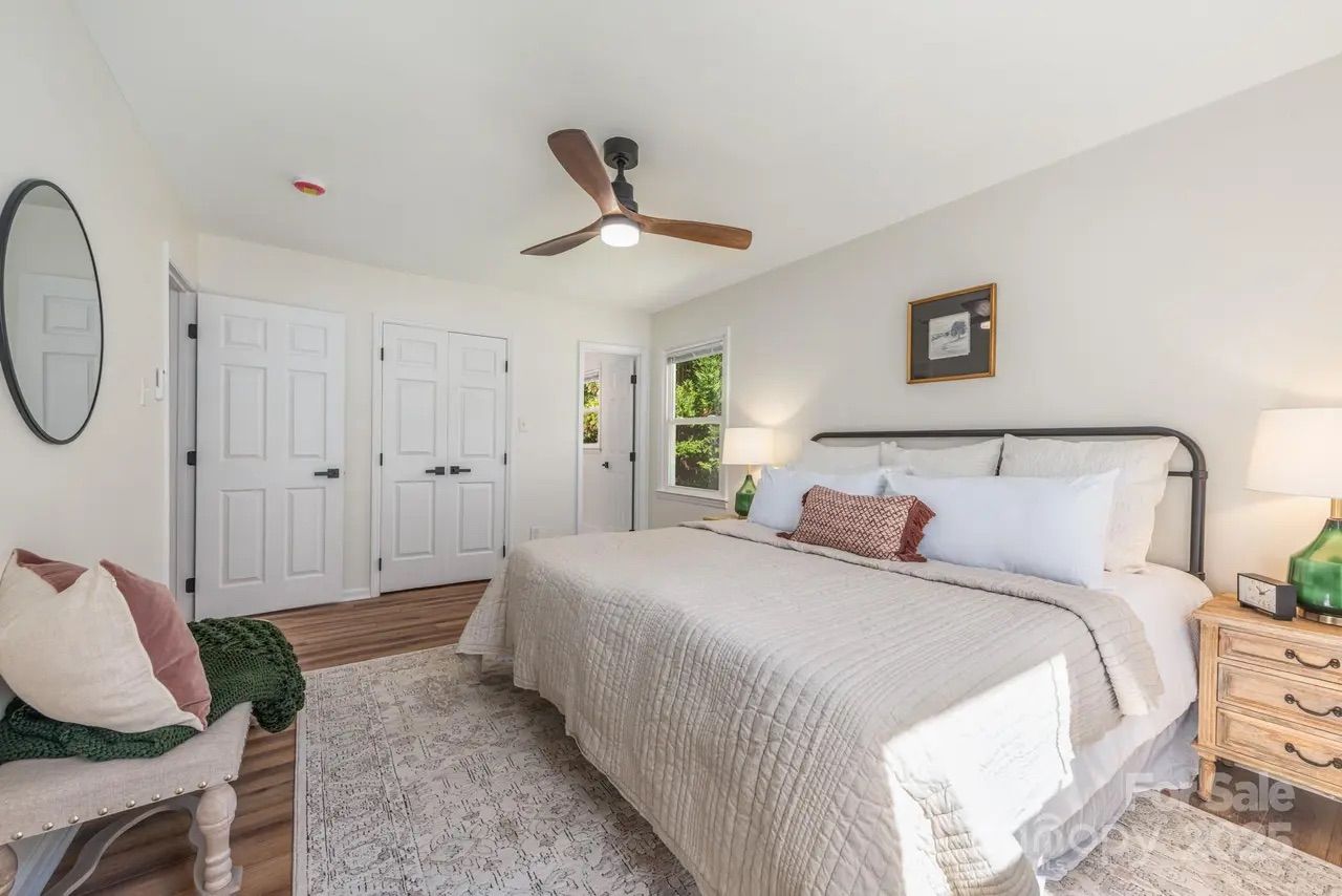 Bright bedroom with a queen bed dressed in beige quilt, white pillows, wooden nightstand with green lamp and clock, round mirror, wooden bench with pillows, ceiling fan, and three white doors.