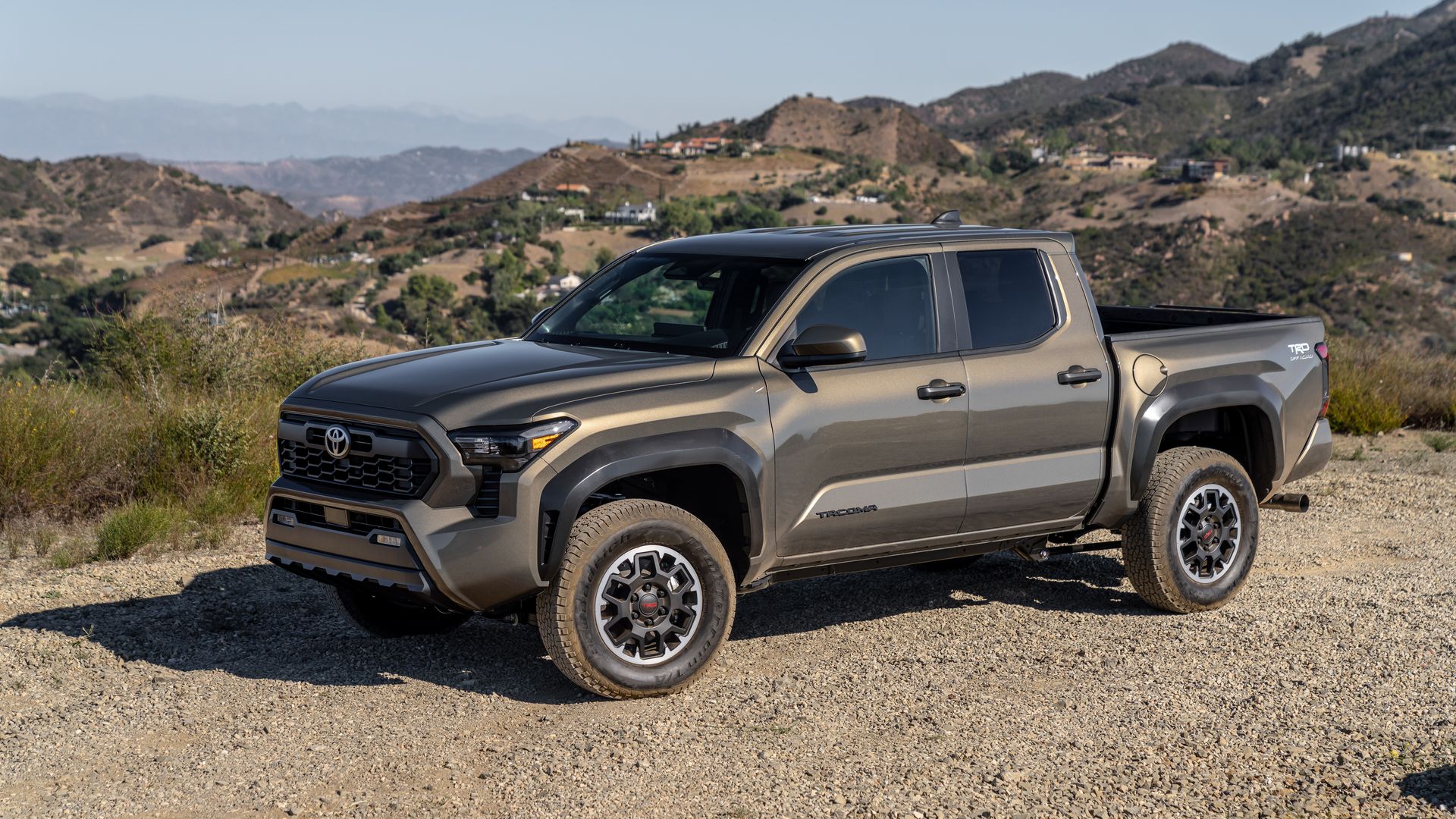 A gray Toyota Tacoma pickup truck parked on a rocky dirt road, with dry, rolling hills and scattered houses in the background.