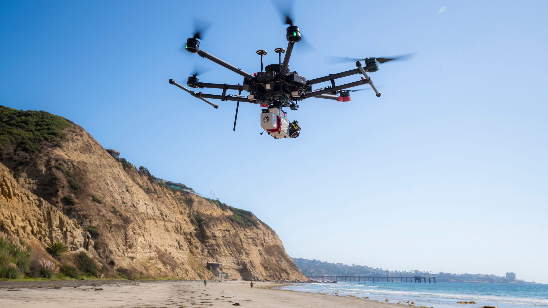A drone with camera flying over a sandy beach with cliffs and ocean under clear blue sky.