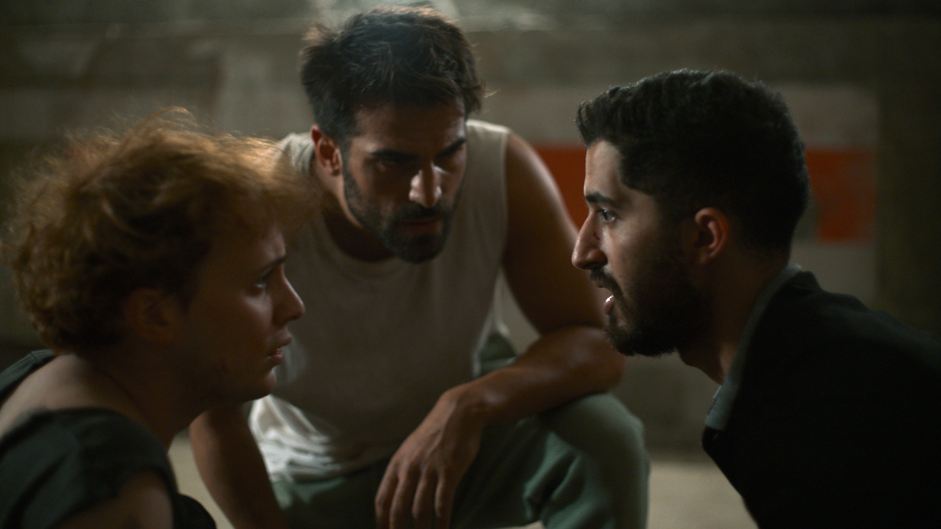 Three people with serious expressions are huddled together in a dimly lit room.