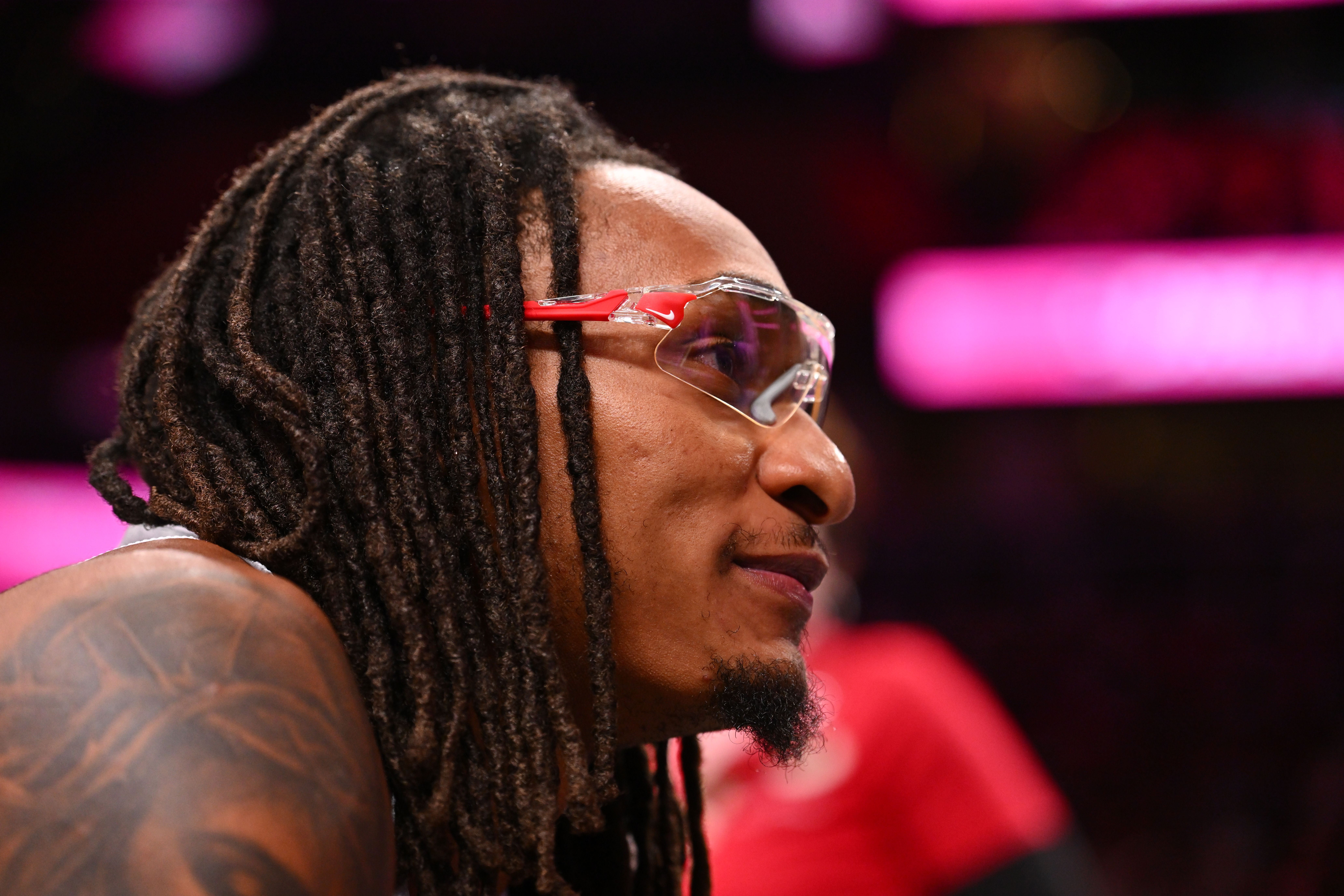 Close-up profile of a person with long black dreadlocks wearing clear protective glasses with red temples, against pink stage lighting.