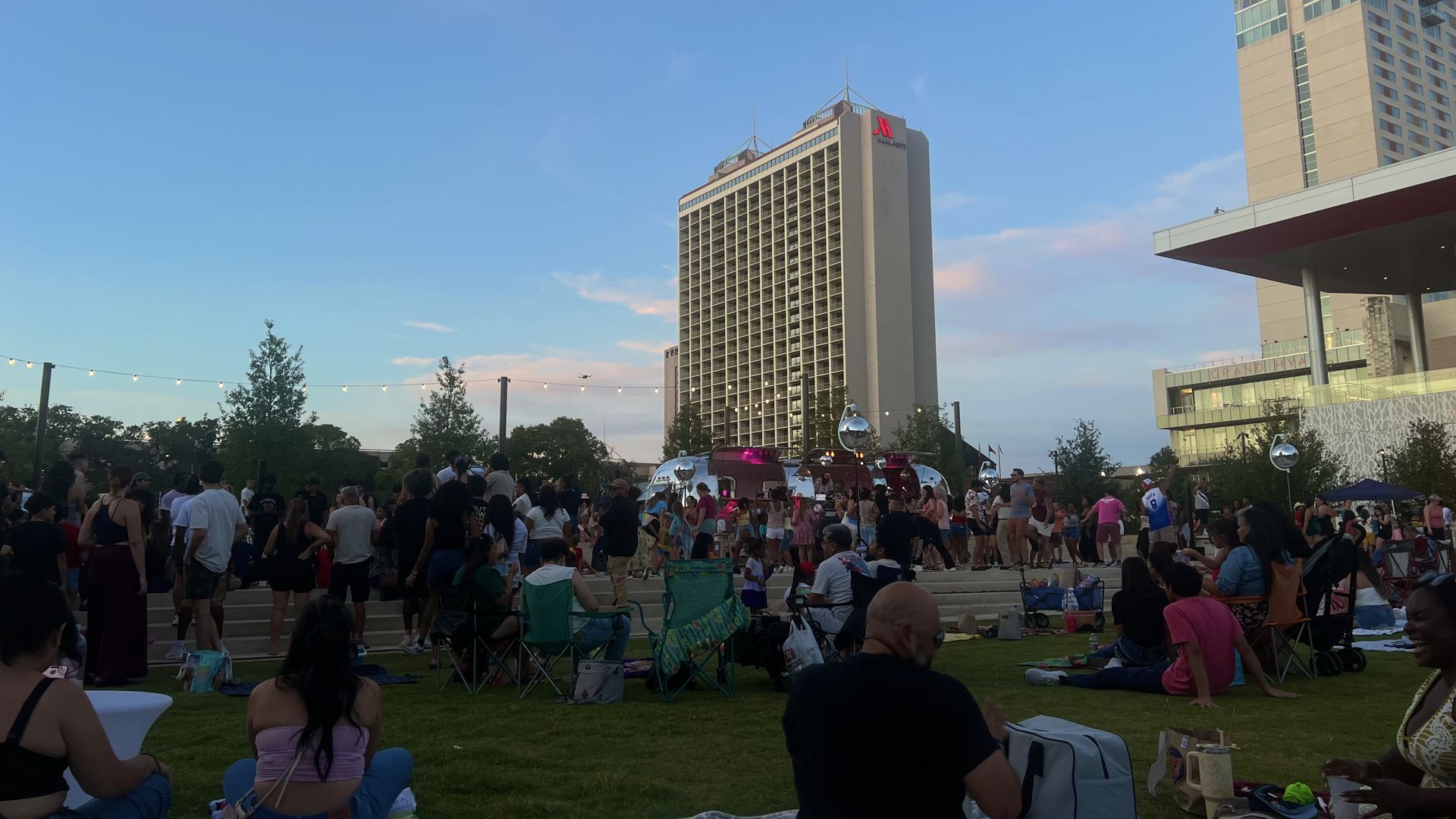 Outdoor evening event with people sitting on grass and standing near a large building with Marriott logo, string lights overhead, clear blue sky with pink clouds, and trees around.