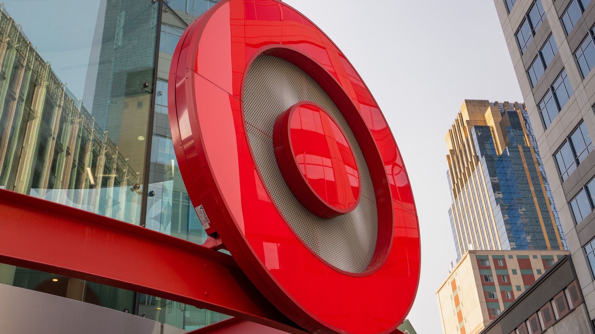 Large red circular Target logo mounted on a red beam outside a building, with skyscrapers and a clear sky in the background.