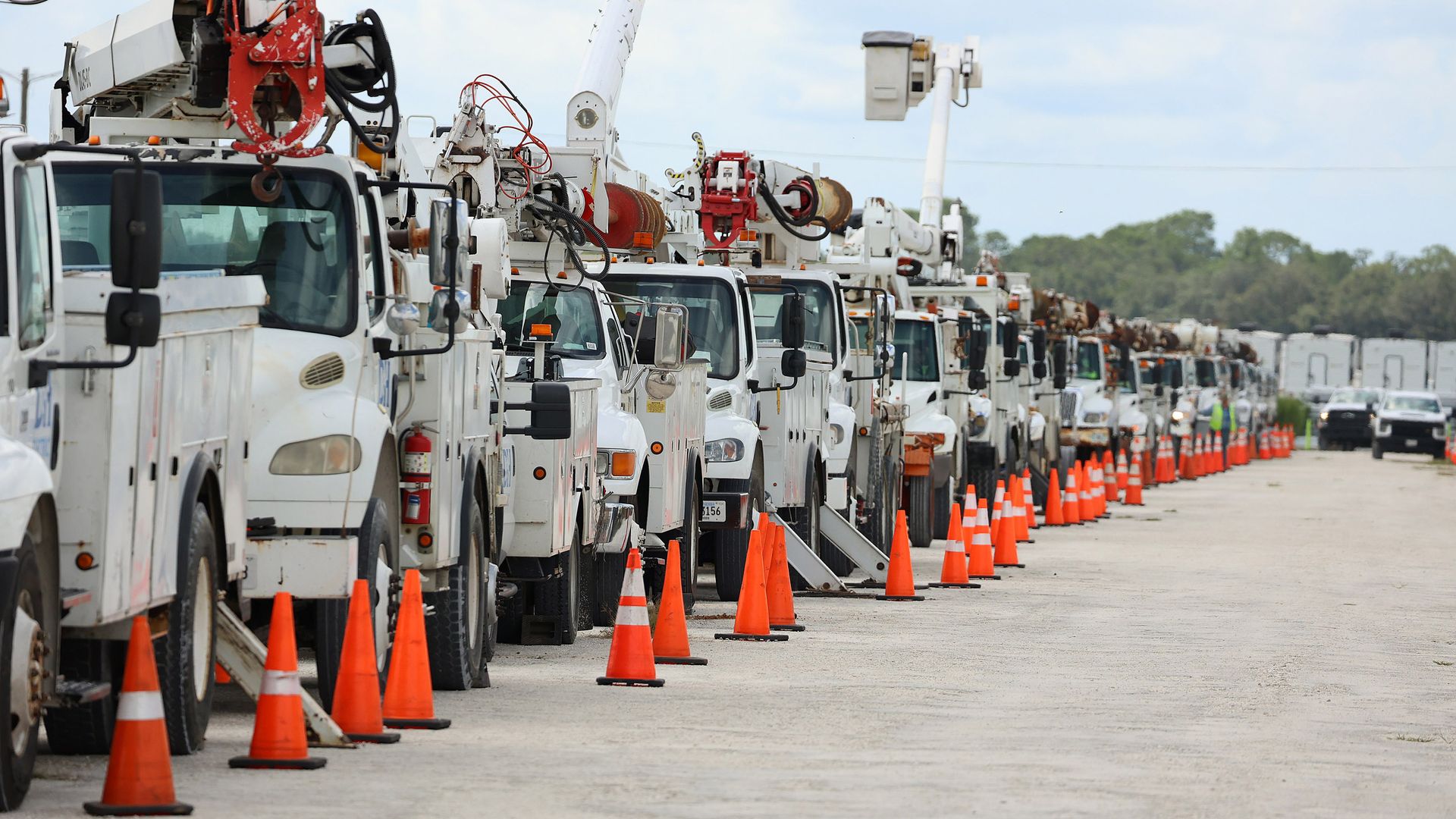 A line of white bucket trucks cordoned off on a dirt road with orange traffic cones. 