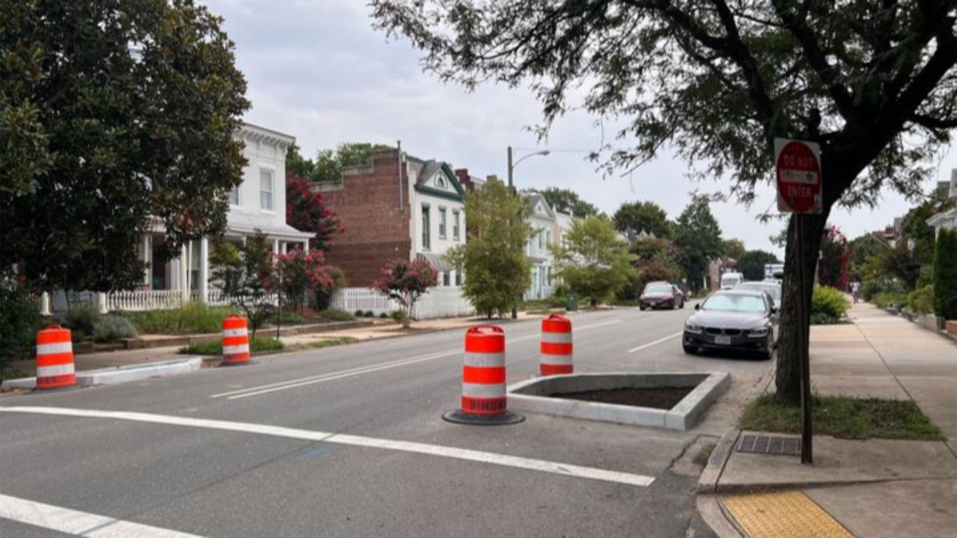 Street view with orange construction barrels around a rectangular concrete planter box on a quiet neighborhood street lined with houses, trees, and parked cars under a cloudy sky.