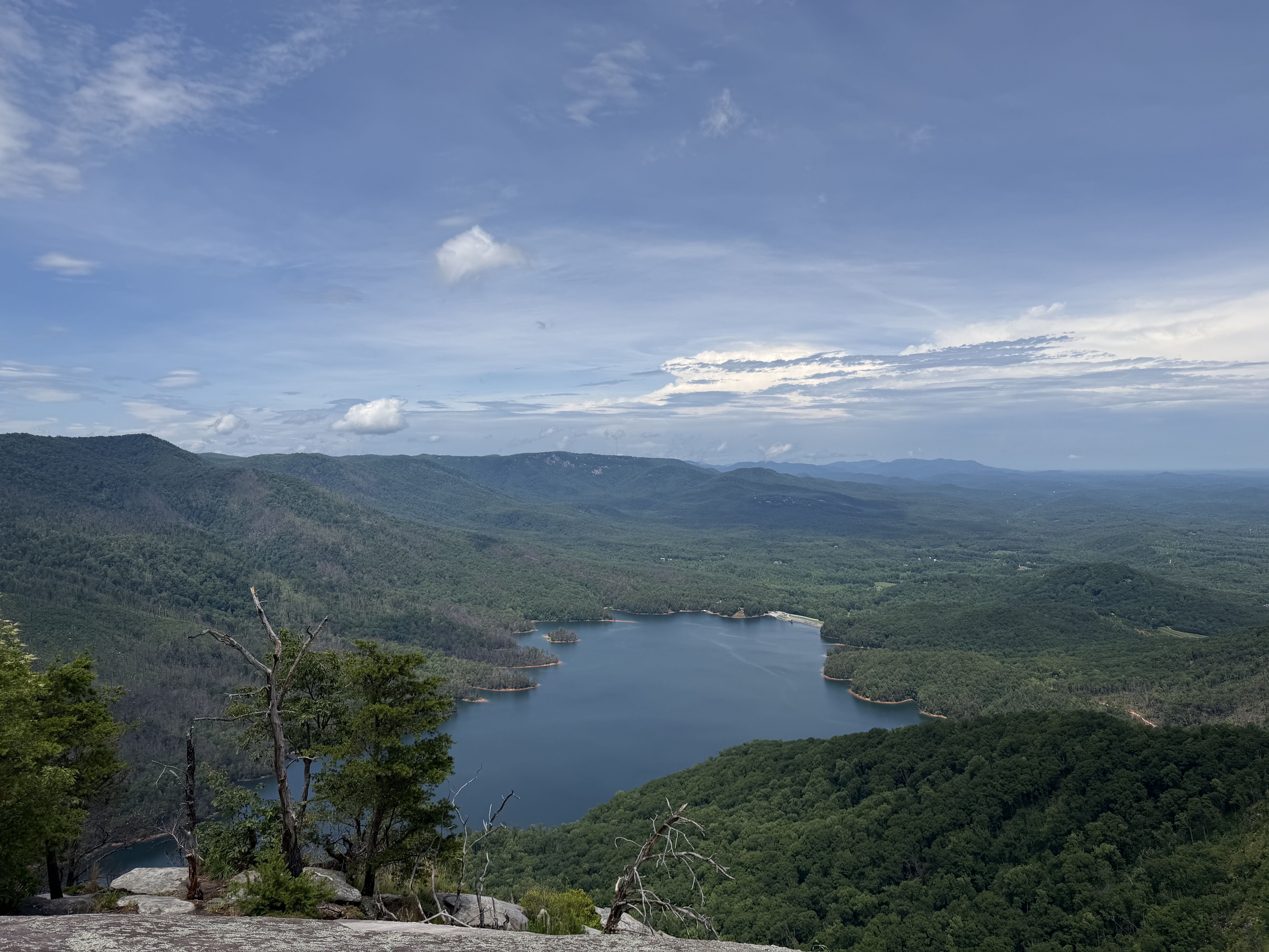 Wide view of a blue lake and green rolling hills under a bright blue sky with scattered clouds; foreground shows rocky ledge and sparse trees, framing the landscape.