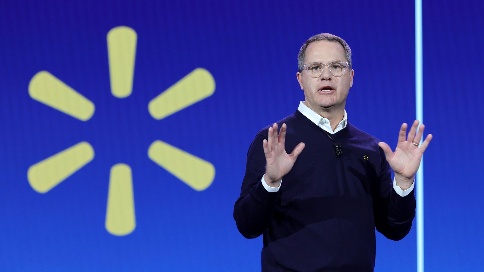 A man wearing a dark sweater is speaking on stage with a blue background and large yellow Walmart logo behind him.