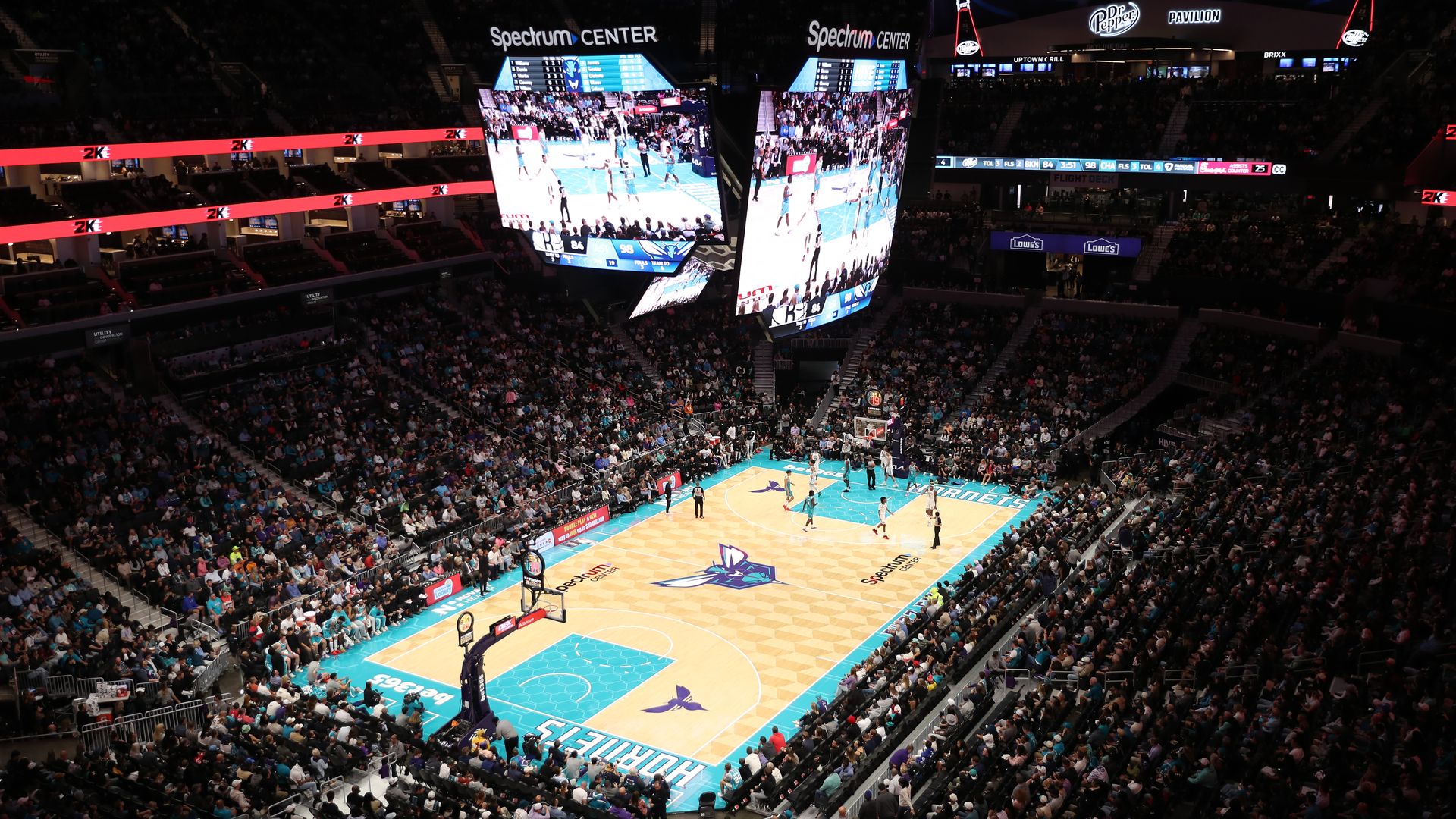 A packed basketball arena with a game in progress on a court marked "Hornets" in teal and purple, with large screens hanging from the ceiling and brands like Spectrum and Dr Pepper visible.