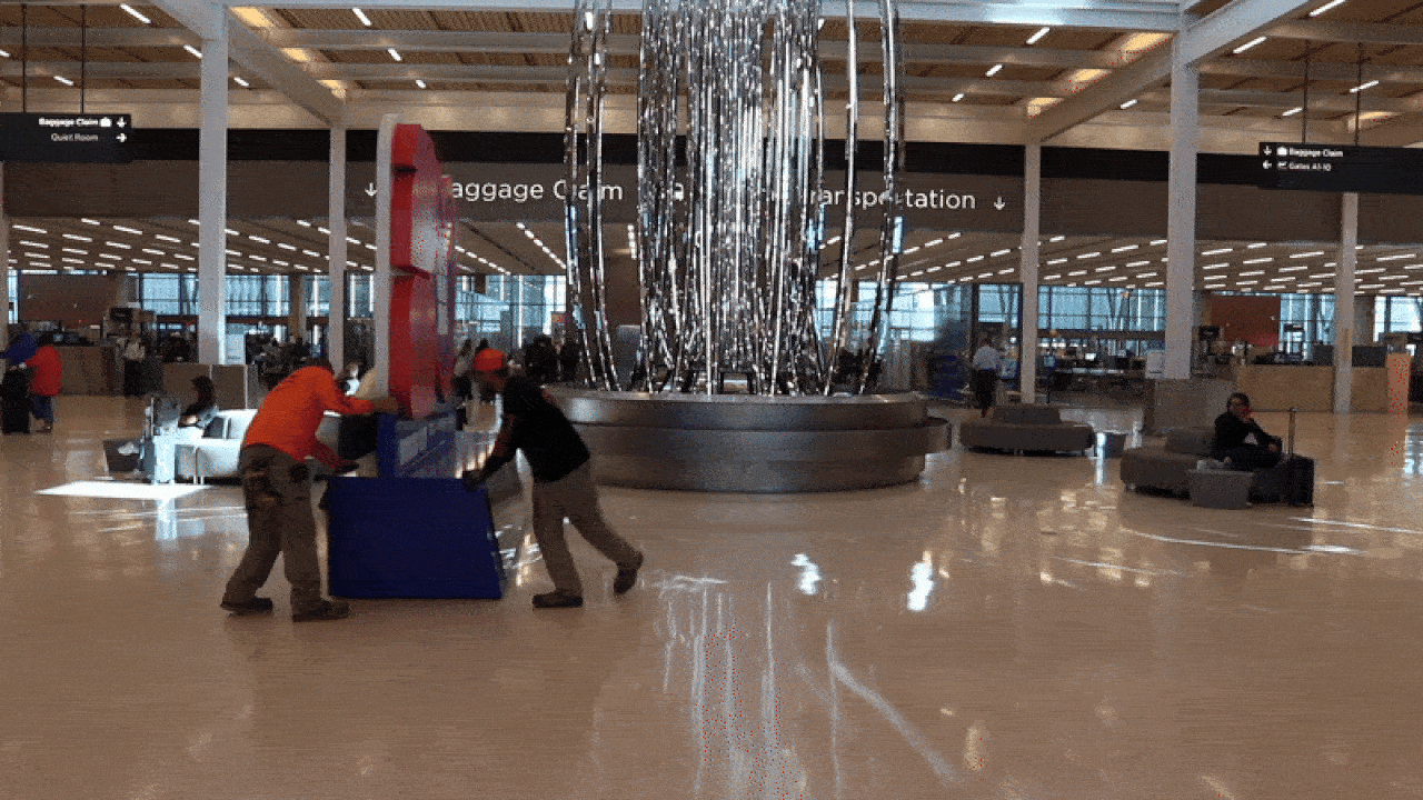 Two airport workers in orange and black move a large blue sign near a silver art installation in a bright, spacious terminal with signage pointing to baggage claim and gates.