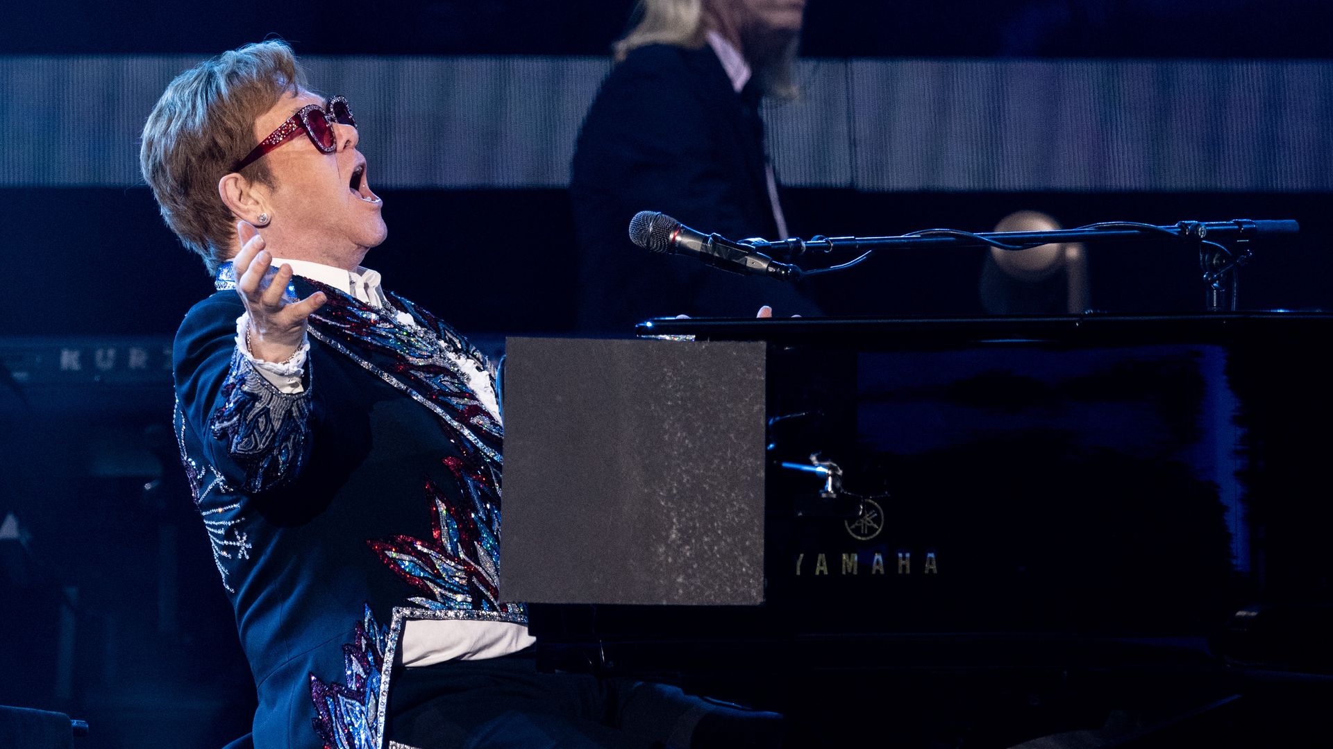 Elton John at a piano singing while wearing a sequined tuxedo and bejeweled glasses