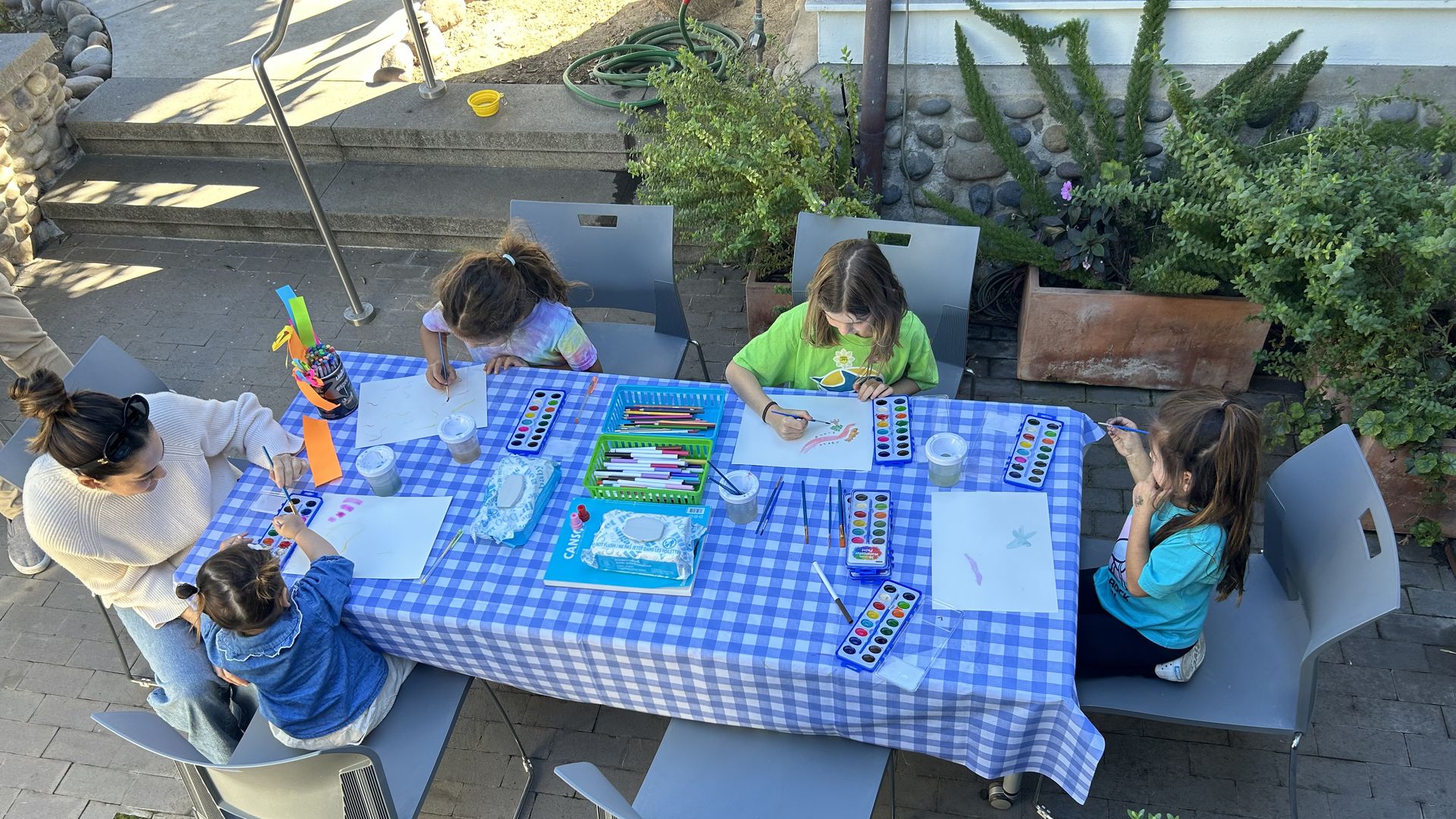 Five people, including children, seated around a table with a blue and white checkered tablecloth, engaging in painting and drawing activities outdoors with watercolor sets and art supplies.