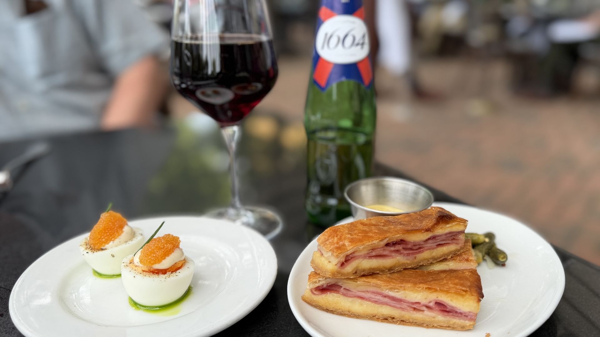 Outdoor cafe table with a glass of red wine, a green bottle labeled LAGER 1664, and two plates: deviled eggs with orange roe and ham-filled puff pastry slices; blurred diners in the background.