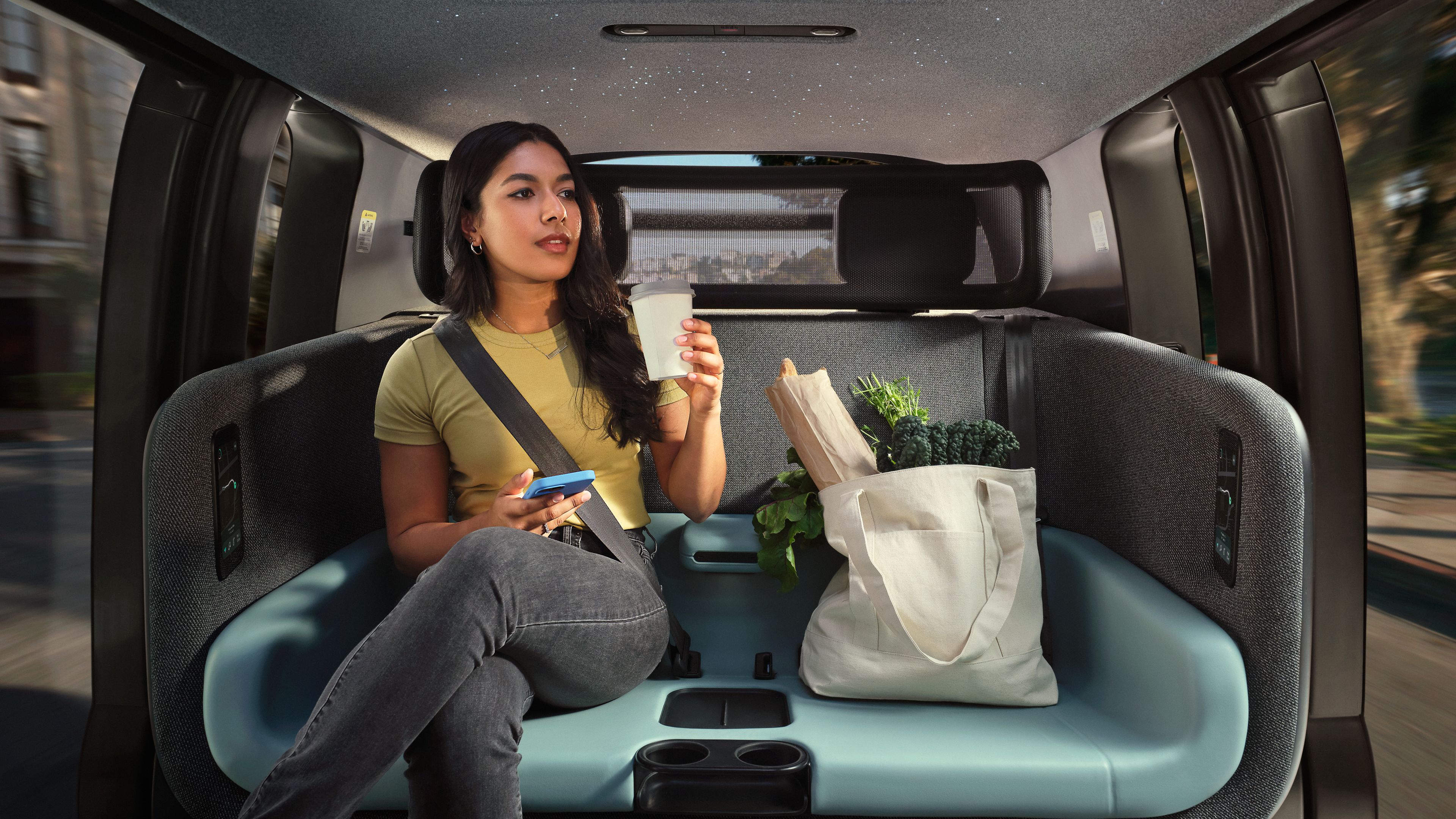 Woman in yellow shirt, gray jeans, sitting in backseat of modern vehicle holding cup and phone, next to a tote bag with groceries including greens and baguette, seatbelt fastened.