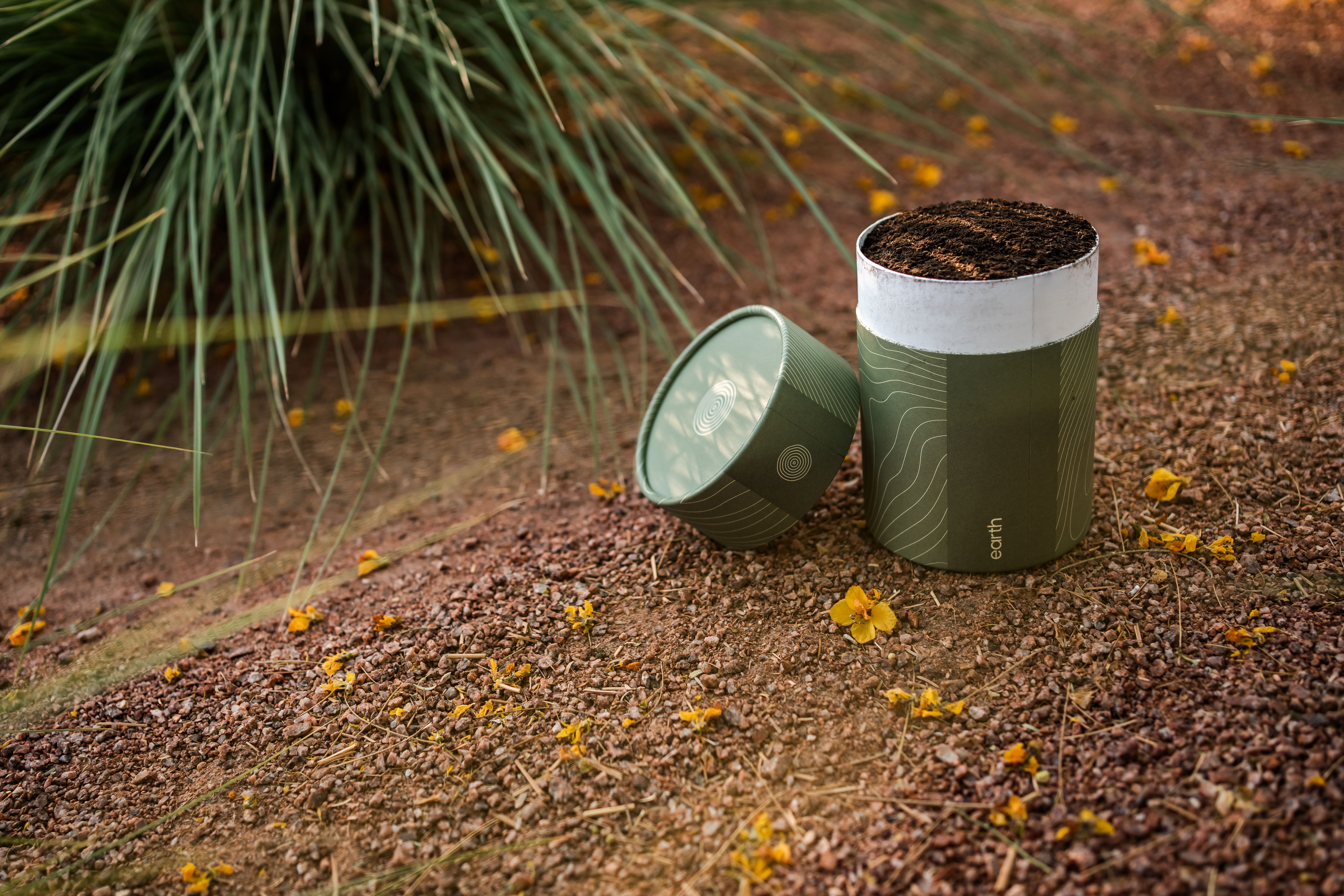 A green cylindrical compost pot filled with dark soil sits on a gravel path outdoors, with its lid leaning nearby. Yellow flowers litter the ground, and tall grasses shade the scene.