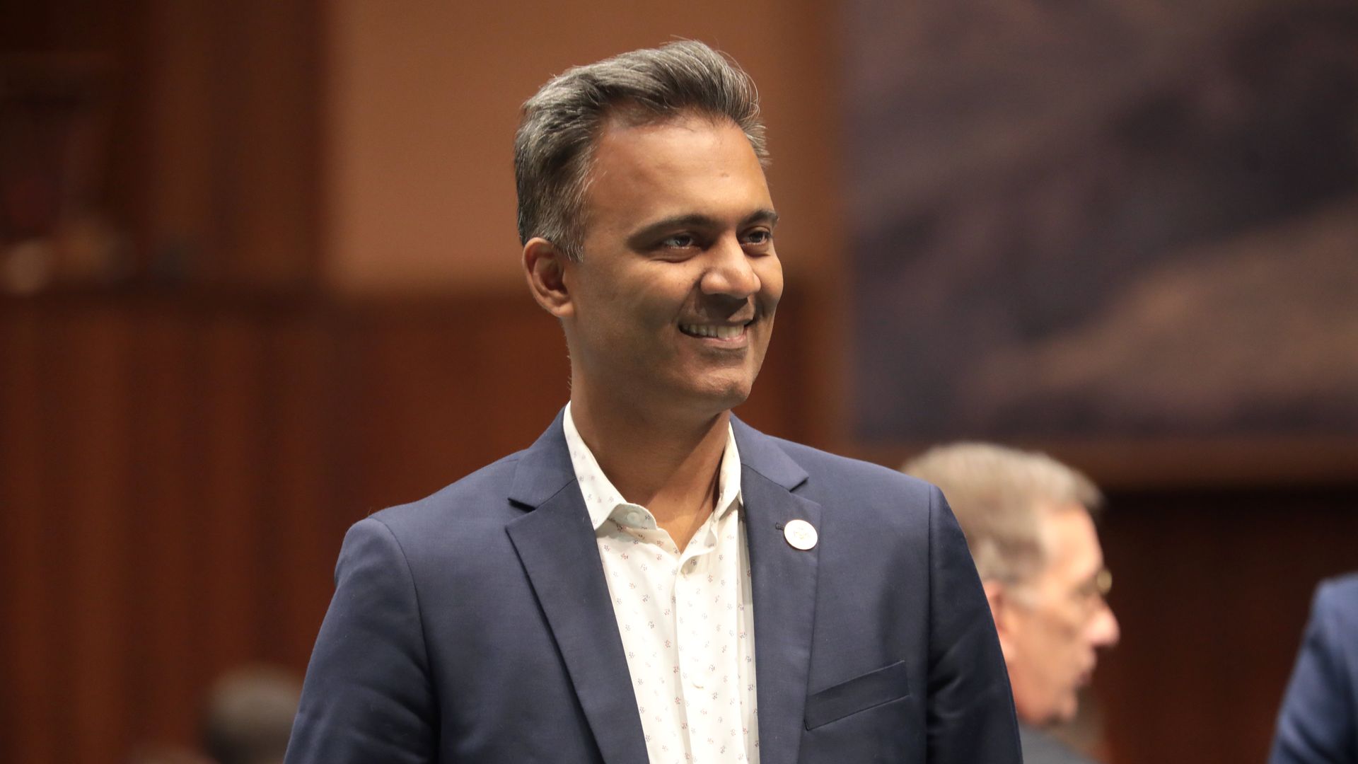 Amish Shah with gray hair wearing a navy blazer and a white patterned shirt smiles at an indoor event, with a blurred crowd and a warm brown background.