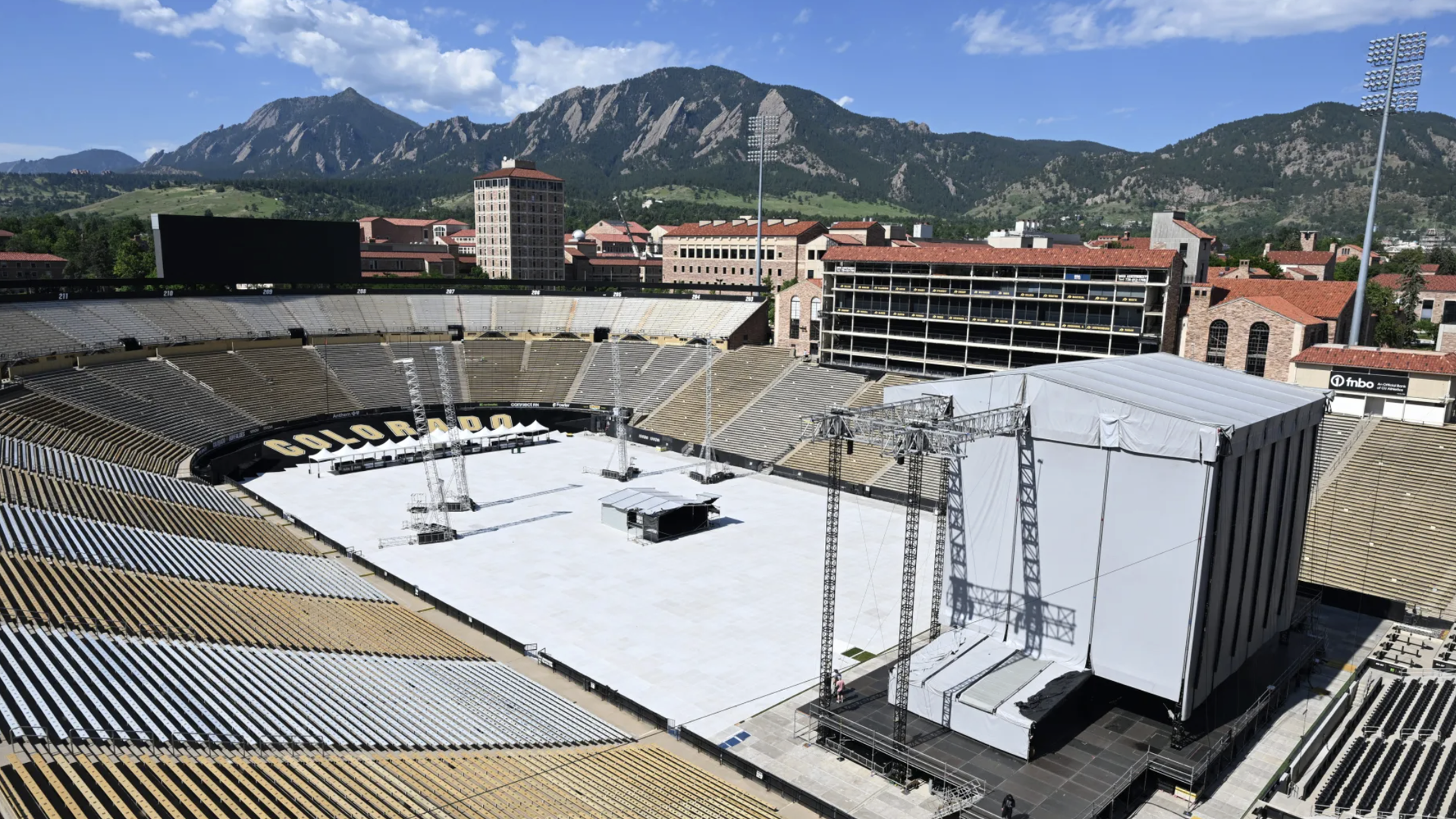Empty Colorado Folsom Field with beige and gray seats, large white stage covered with canopy, black and yellow letters spelling COLORADO, and mountains under a partly cloudy blue sky.