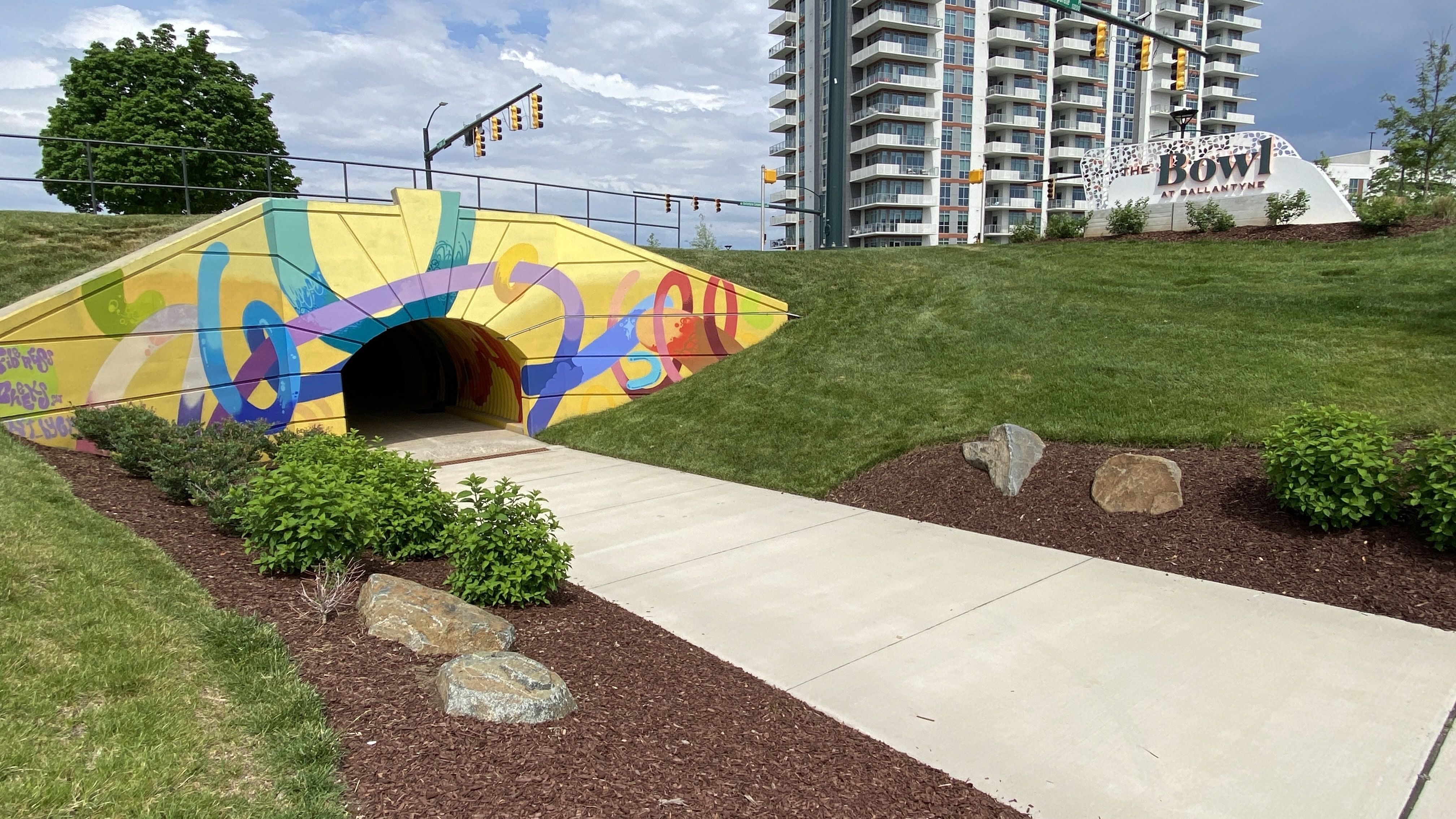 Brightly painted pedestrian tunnel with abstract colorful designs on yellow walls, a concrete path leading into it, surrounded by green grass, bushes, rocks, and an apartment building and sign reading "The Bowl at Ballantyne" in the background.