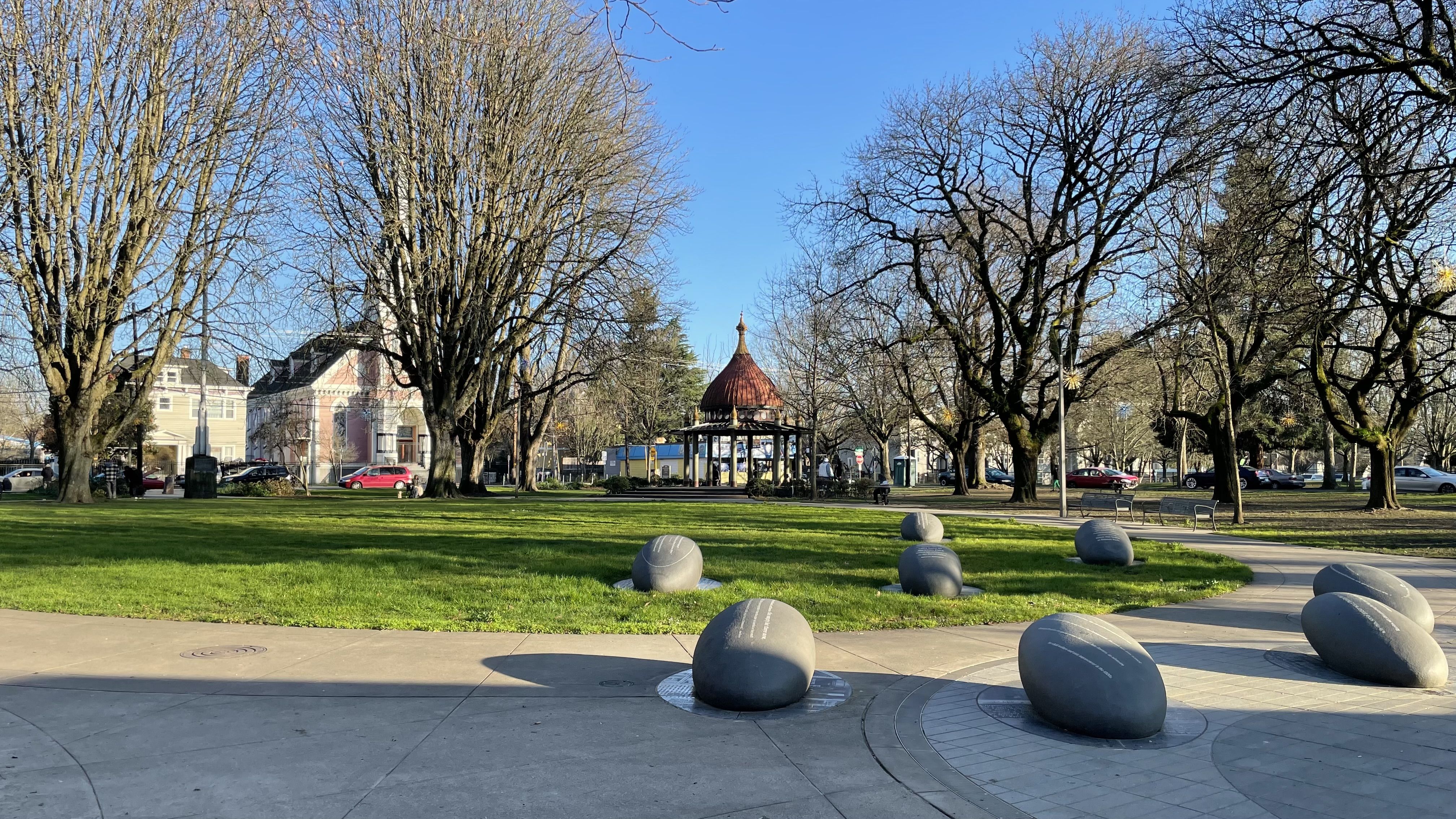 Park scene with large gray stone sculptures on concrete paths, a green grassy area, leafless trees, a small gazebo with a red dome roof, and houses and cars in the background under a clear blue sky.