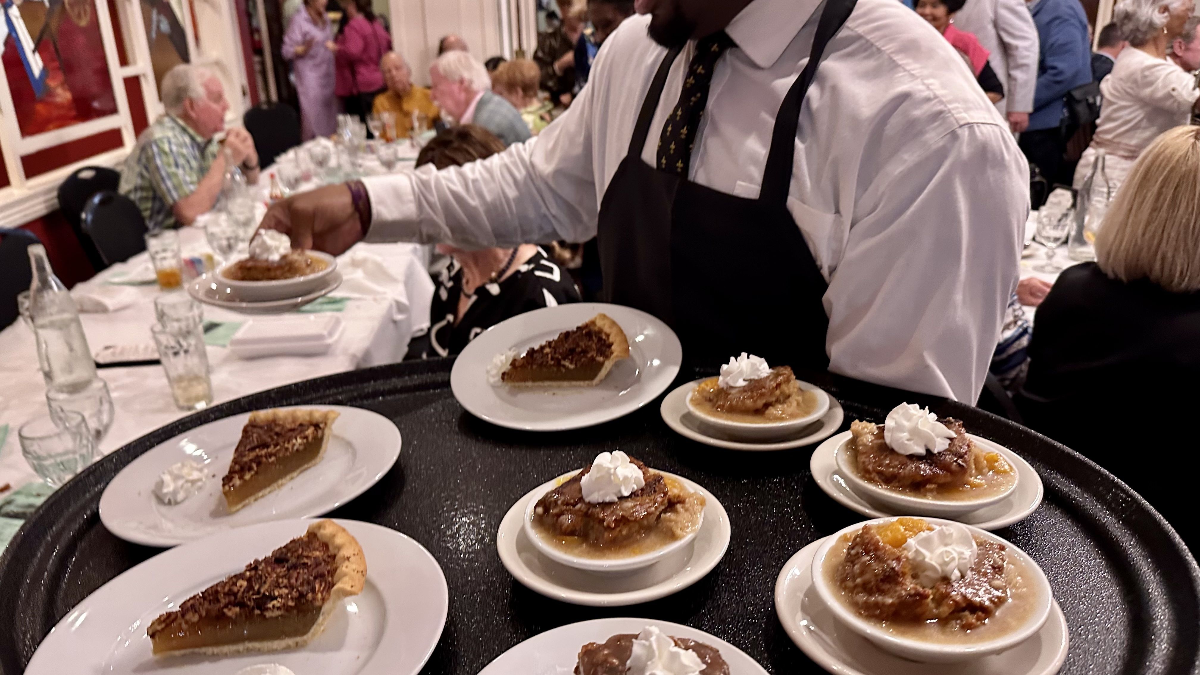 Waiter in a white shirt and black apron carries a large tray of desserts at a crowded banquet: several pie slices and small bowls of cobbler topped with whipped cream.