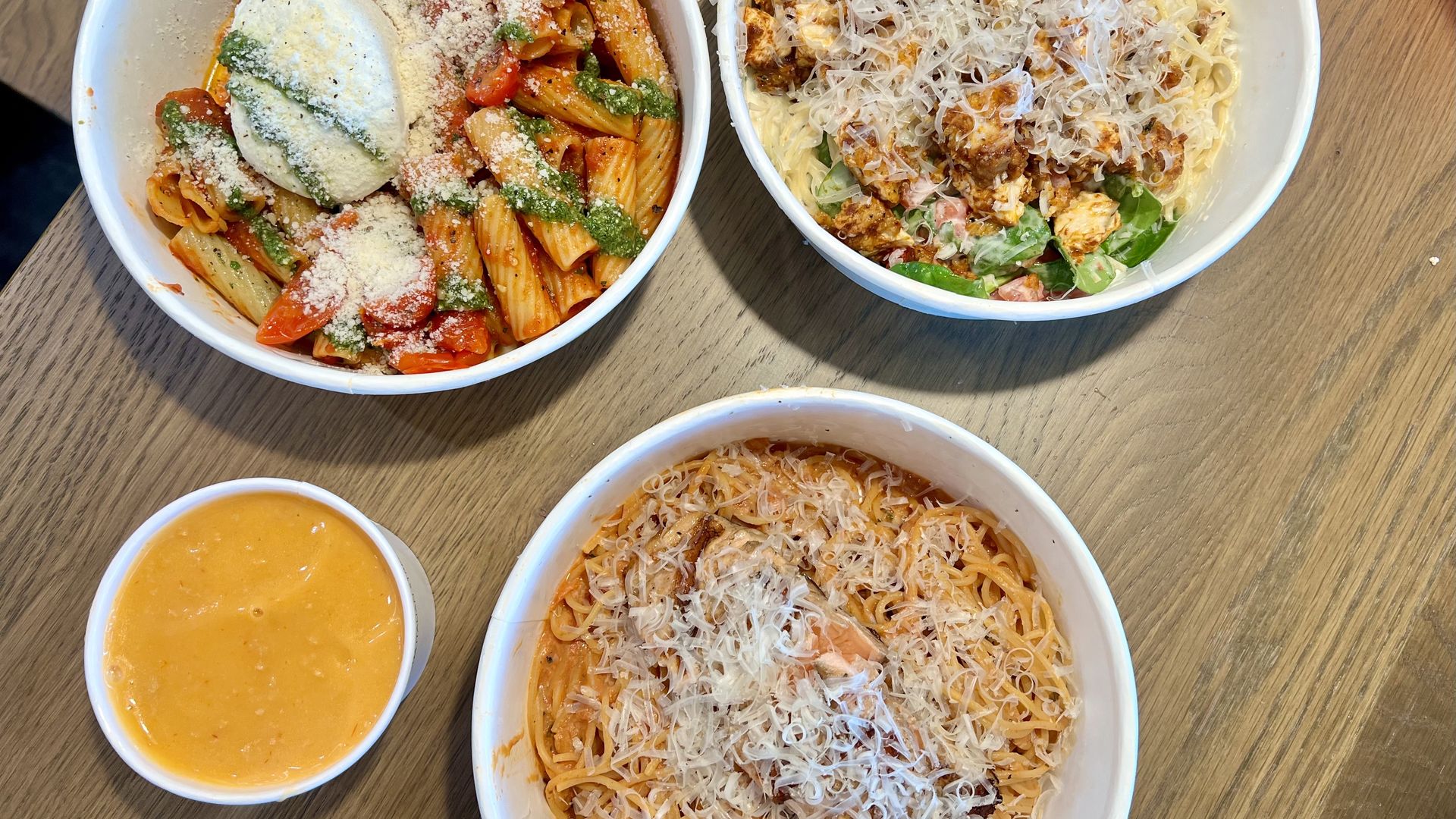 Four bowls of pasta on a wooden table: penne with tomato-vegetable sauce, chicken pasta with cheese, cheese-topped spaghetti; plus a small cup of soup.