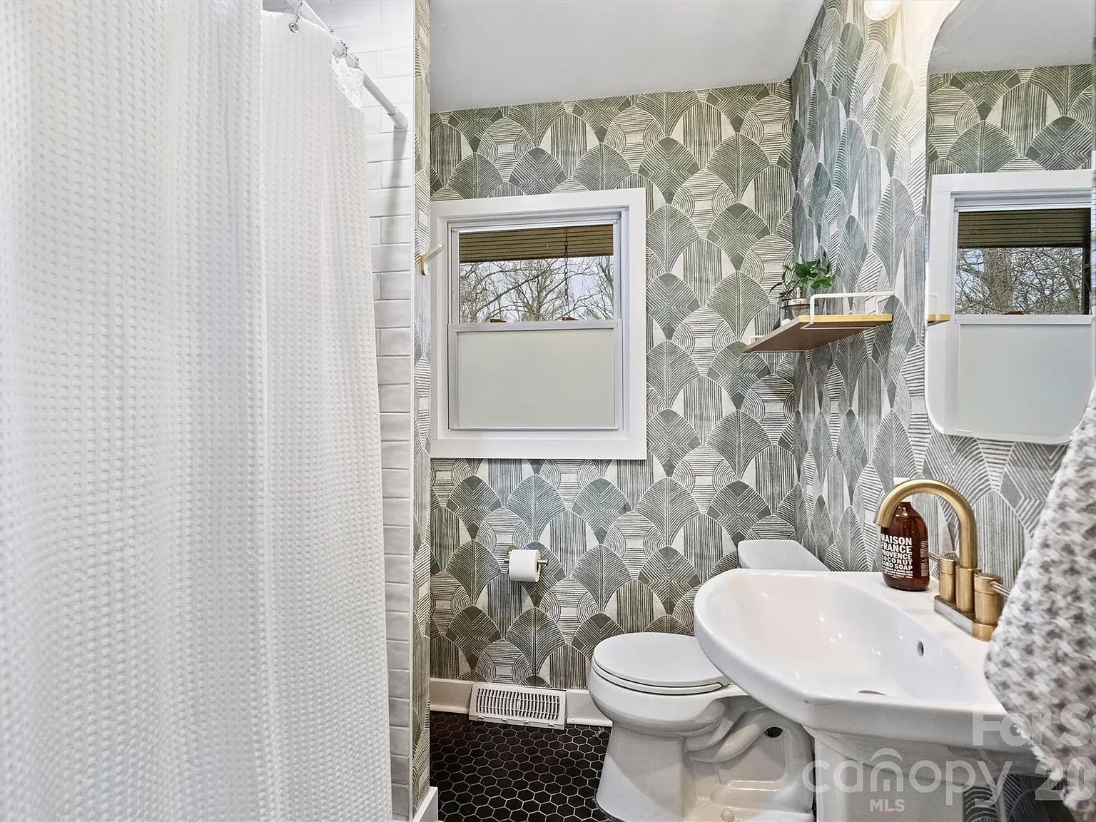 Small bathroom with white fixtures, gold faucet, green and white geometric wallpaper, black hex tile floor, white waffle shower curtain, small window, and floating shelf with plants.