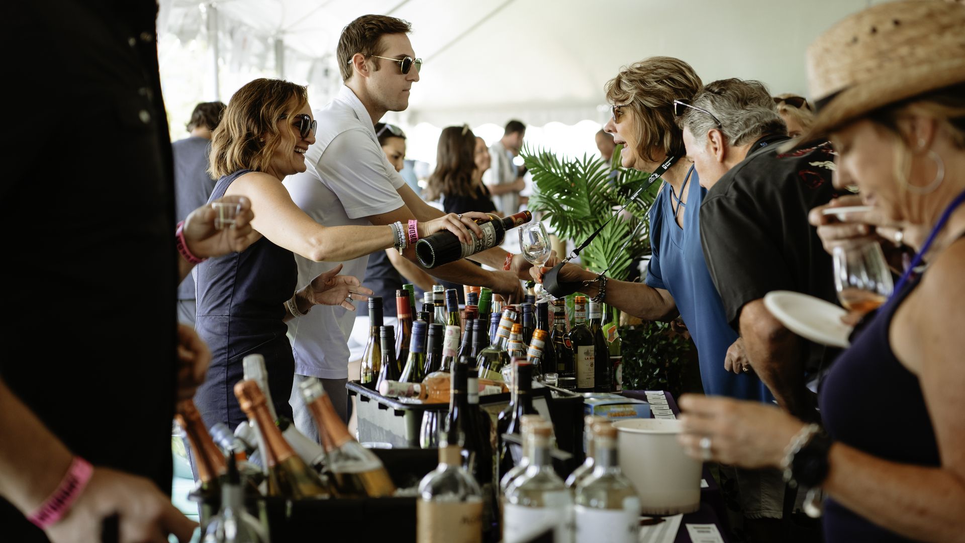 A woman smiling while wearing sunglasses pours wine while another woman holds a glass to take it.