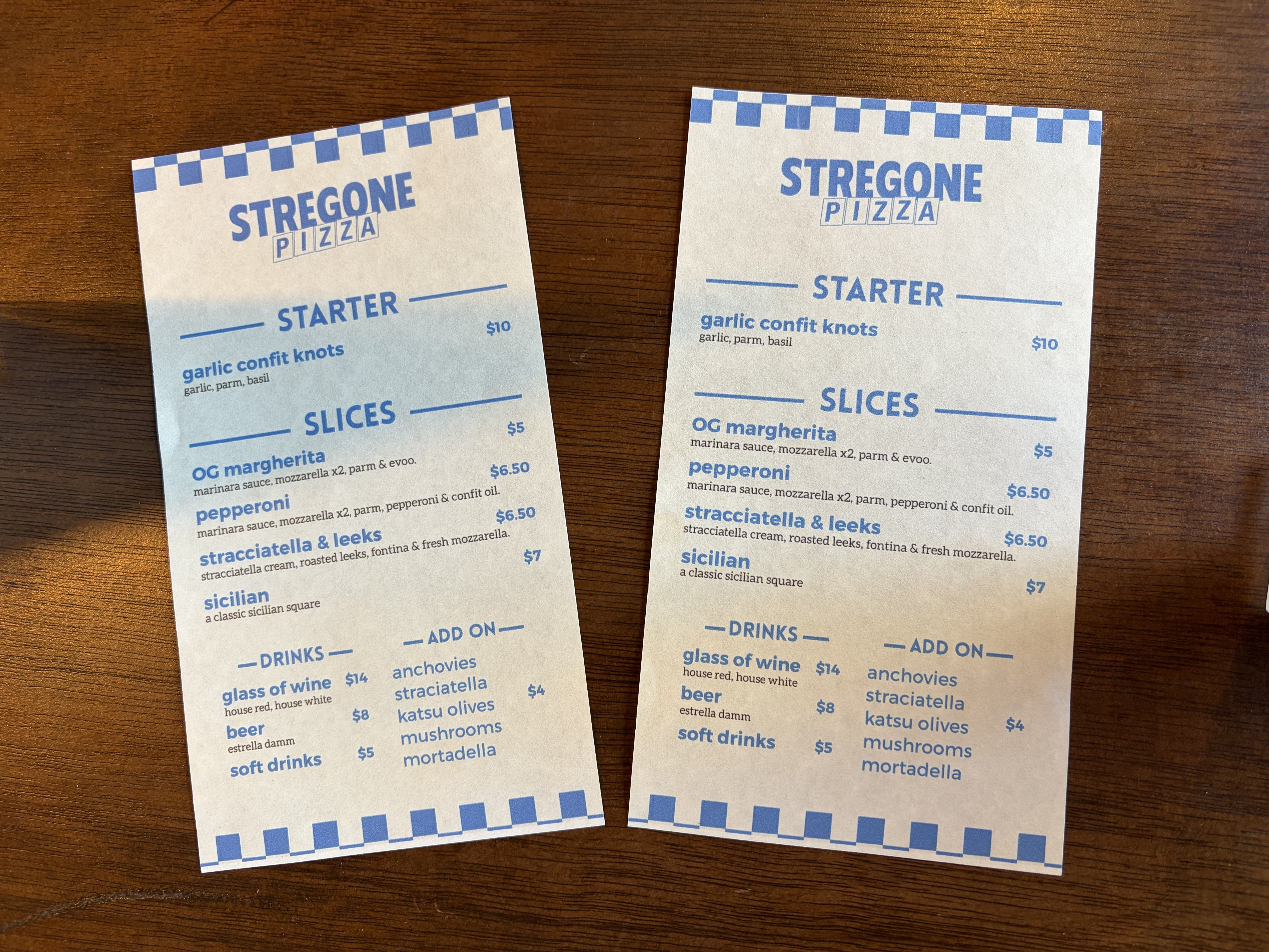 Two pizza menus with blue text and a checkerboard border on a wooden table; logos read "STREGONE PIZZA" with sections "STARTER" and "SLICES", featuring garlic confit knots.