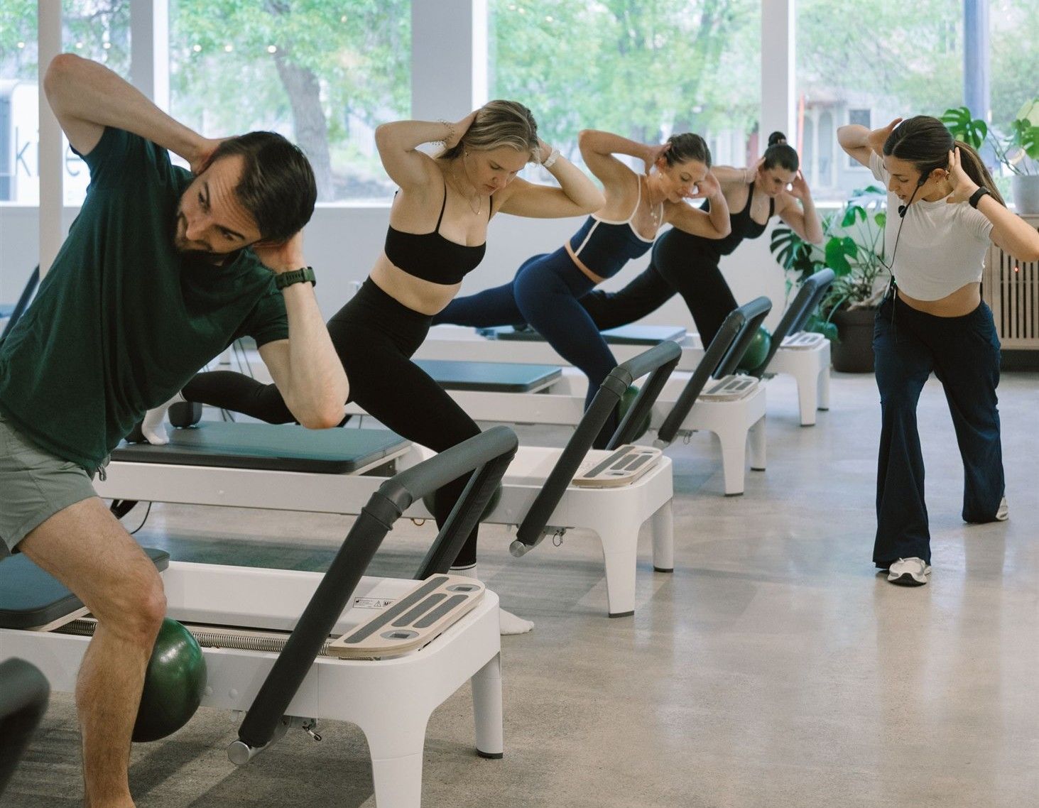 Group fitness class on treadmills in a bright studio; five people bend sideways with hands behind their heads as they exercise, led by a woman with a headset on the right, with plants and windows in back.