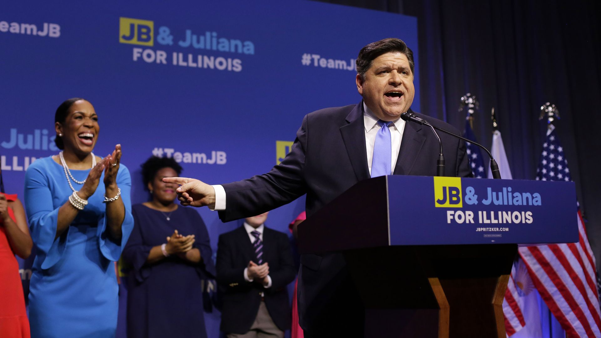 Photo of a man behind a podium gesturing to a woman behind him who is clapping and smiling. 