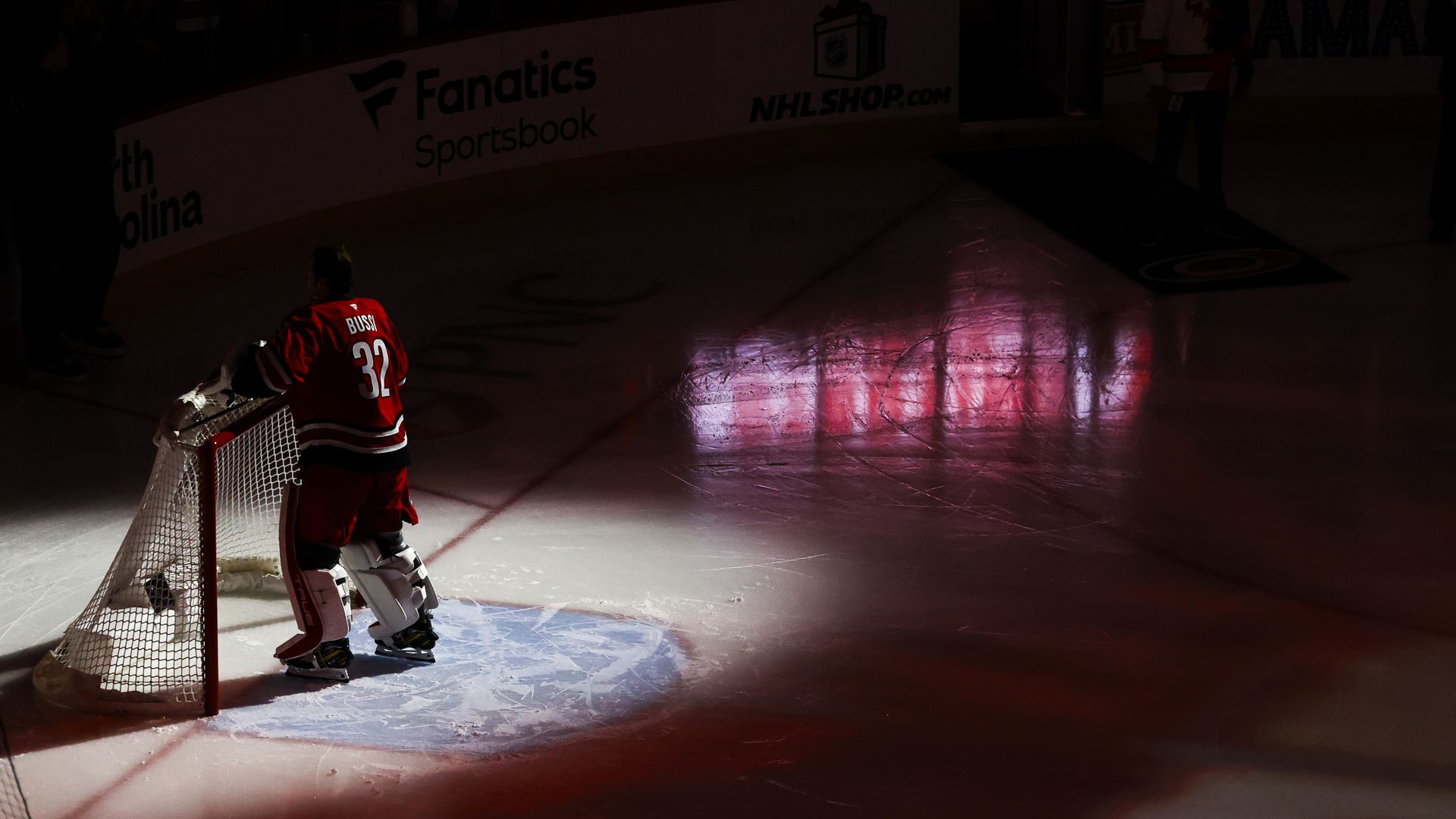 Hockey goalie in red jersey number 32 stands in front of net on ice rink, spotlight on him, dark arena with red lighting reflections on ice.