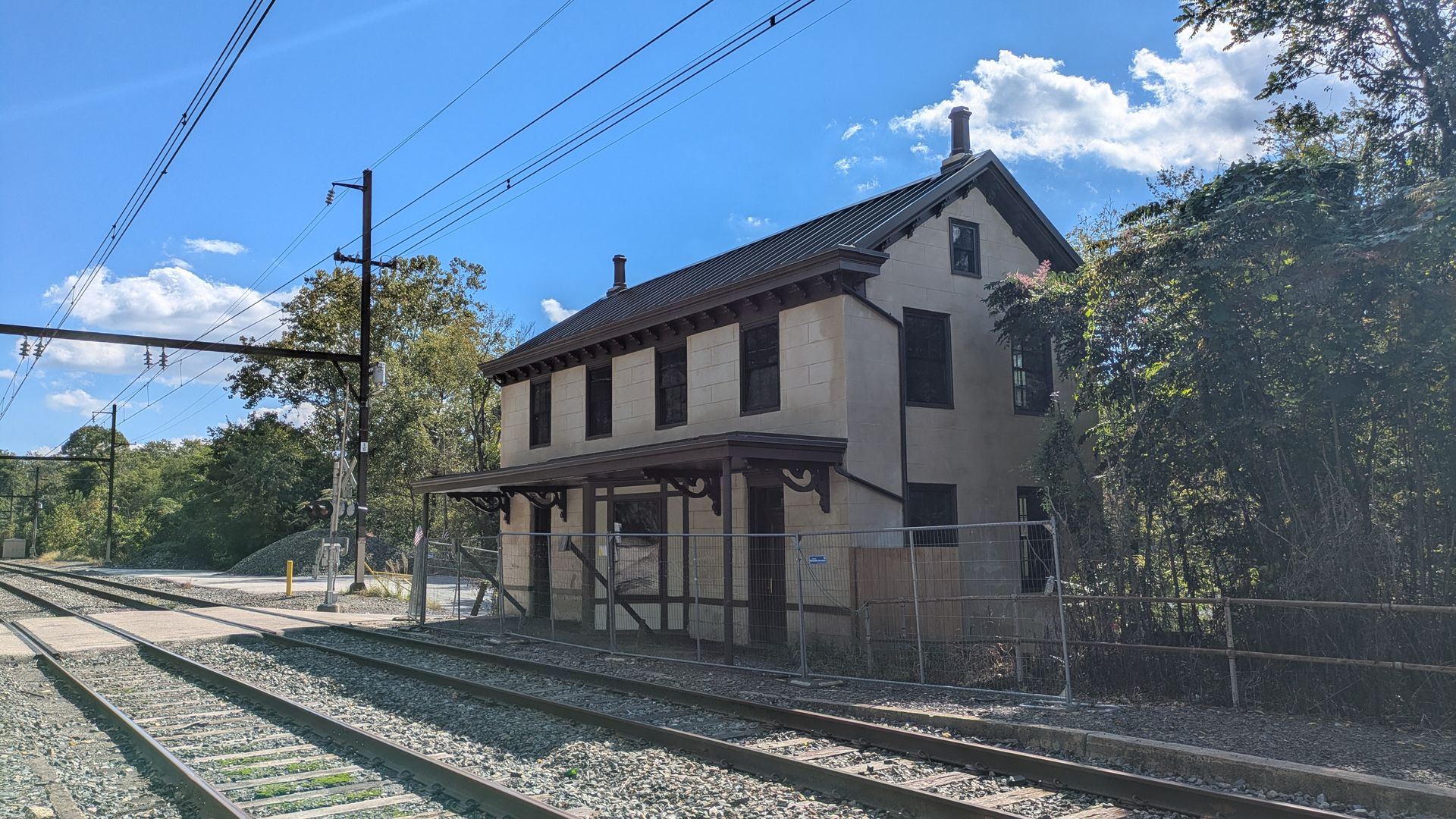Beige two-story building with a dark pitched roof and two chimneys beside train tracks. Overhead power lines cross a bright blue sky, with trees and a chain-link fence nearby.