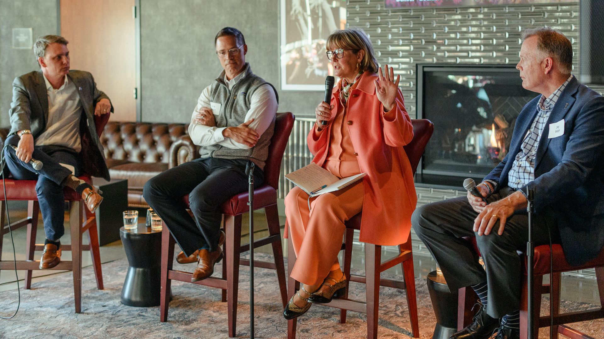 Four panelists seated on stools in a modern lounge; a woman in an orange suit speaks into a microphone as others listen, with a fireplace and a mountain landscape displayed on a screen behind them.