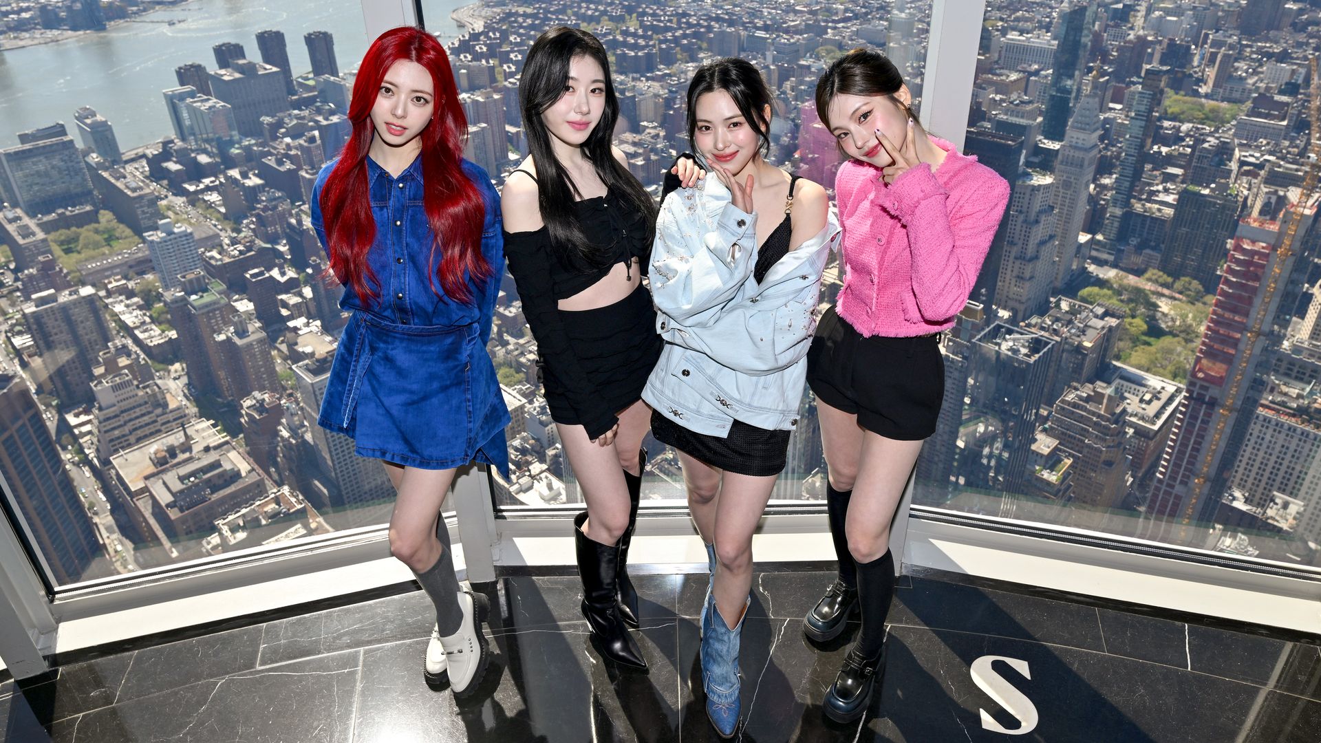 Four women K-Pop stars pose in front plate glass windows offering a panoramic view of Manhattan at the top of a very tall skyscraper