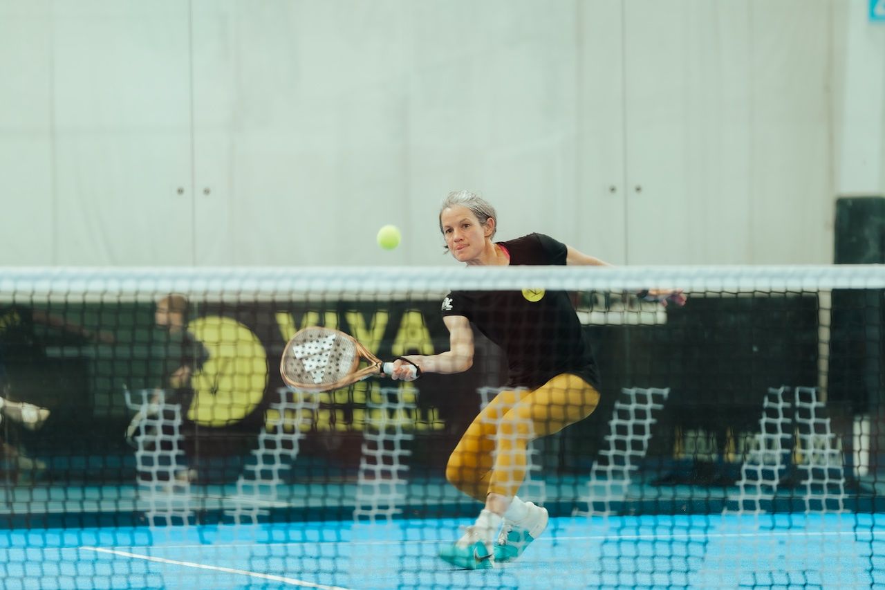 Female padel player on an indoor blue court lunges to hit a ball with a wooden racket, wearing a black top and mustard yellow leggings, white shoes, near the net.
