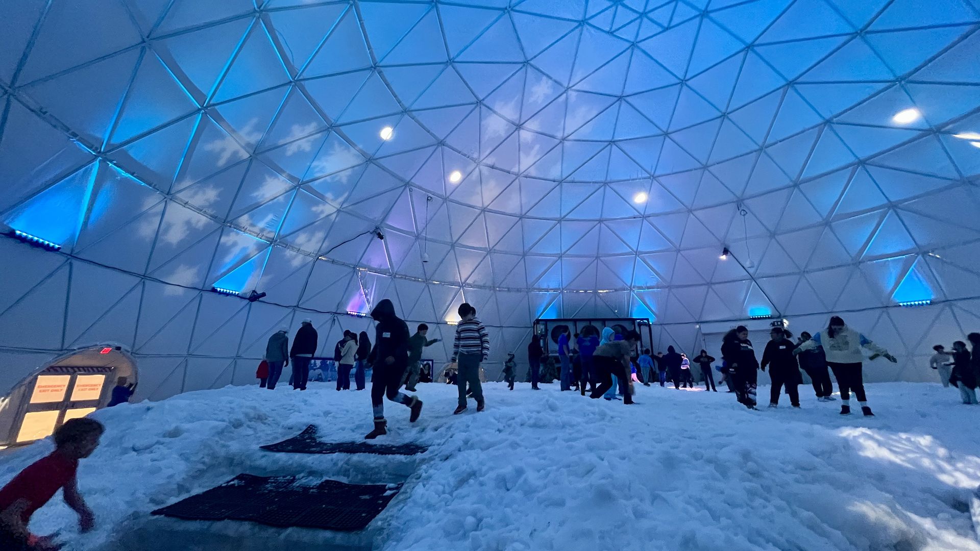 Inside a large geodesic dome with blue and purple lighting, many people are playing and walking on a mound of white snow under a high ceiling with triangular panels.