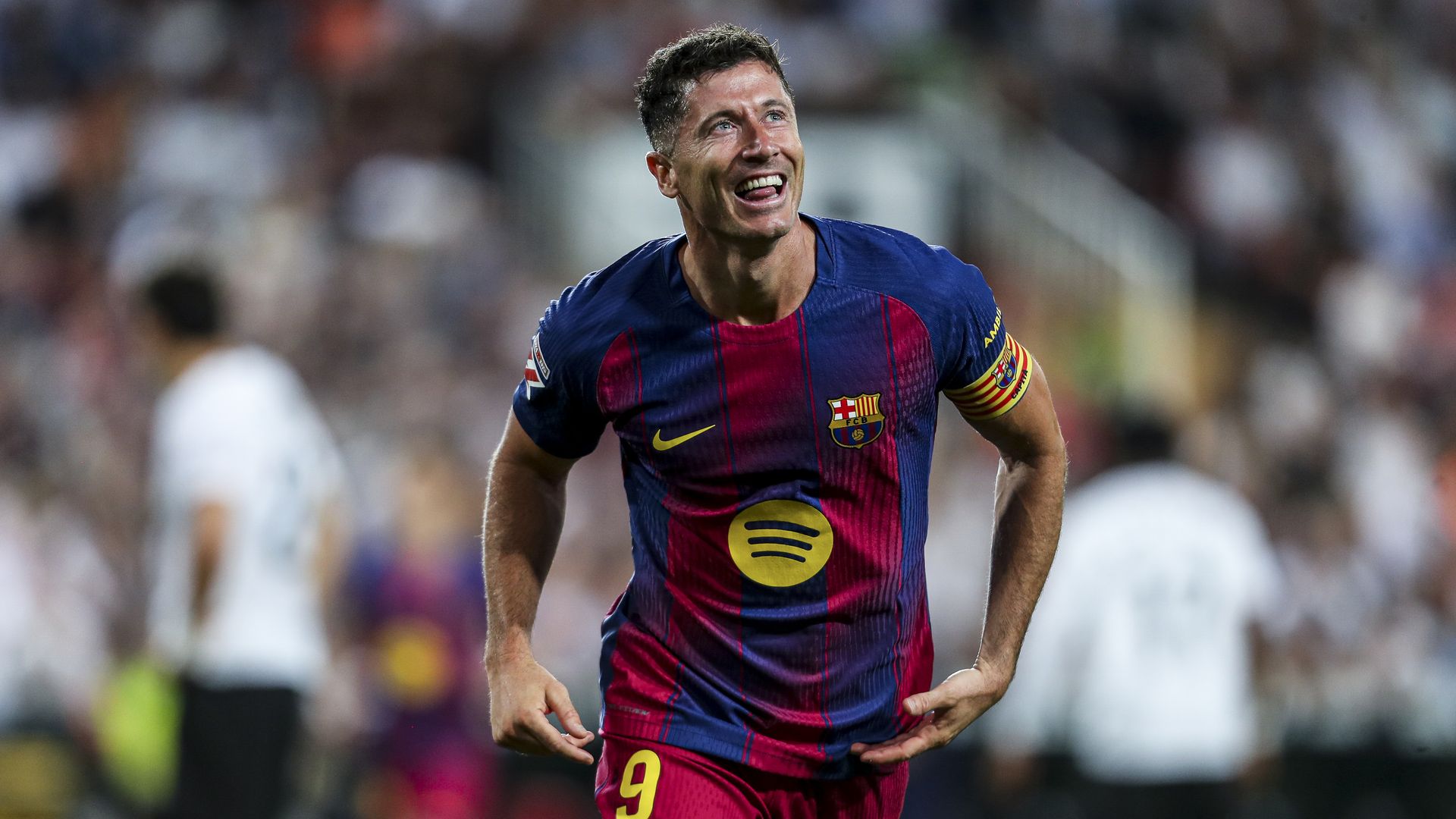 Barcelona soccer player in blue and maroon striped jersey, number 9, smiling while running on the pitch; captain's armband on his left arm; blurred stadium crowd in background.