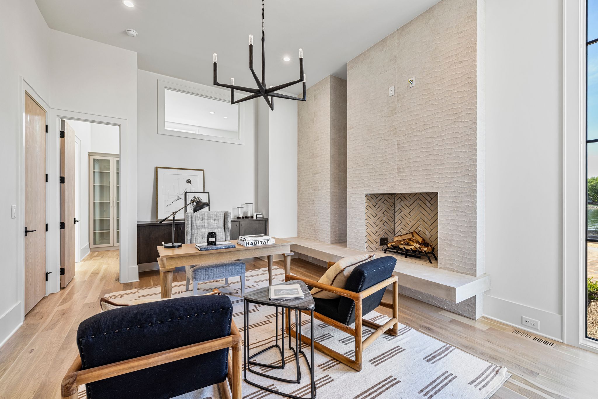 Modern living room with light wood floor, textured beige fireplace, wooden furniture with dark cushions, a desk with books, and large window showing outdoor greenery.
