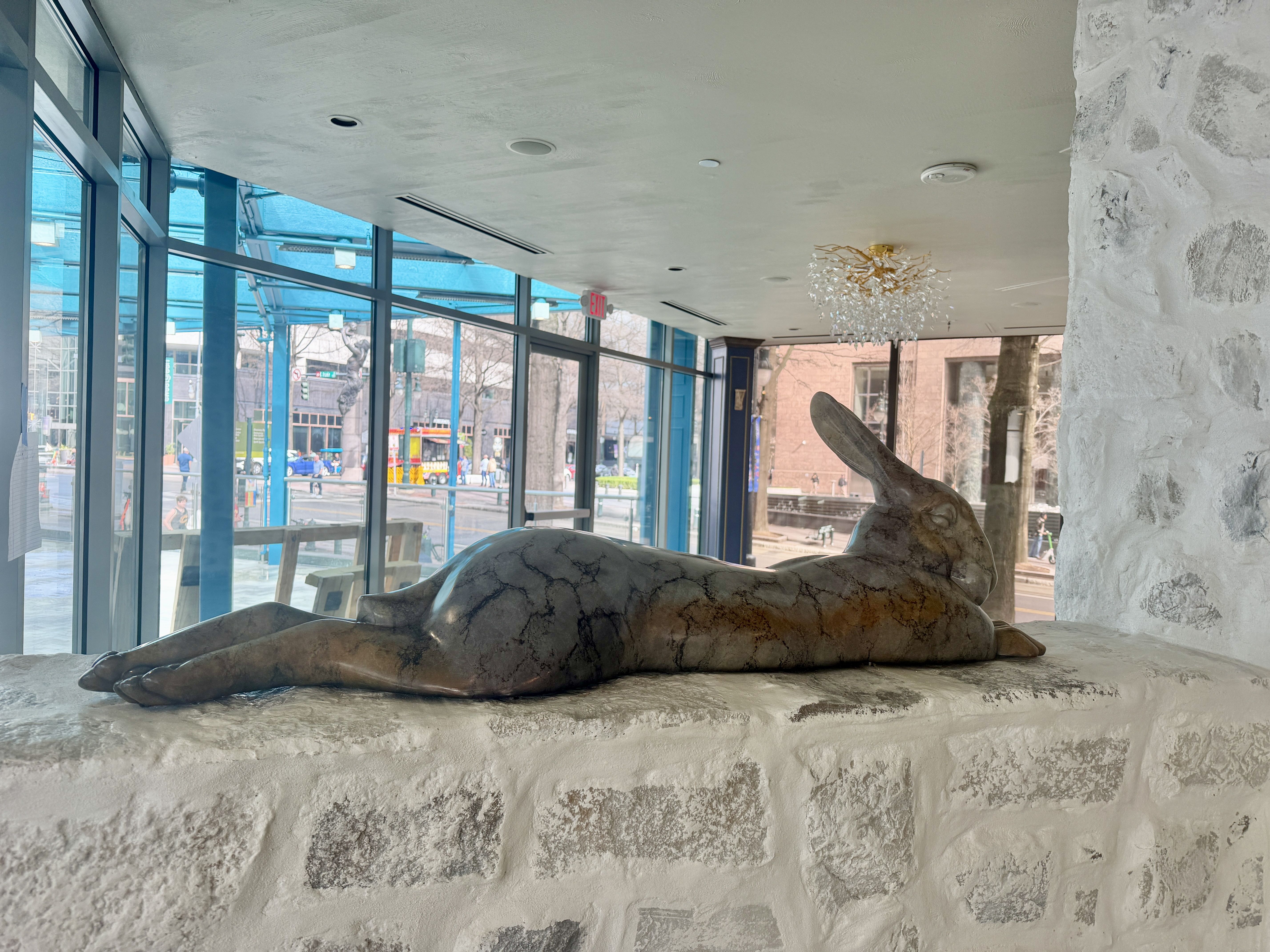 Bronze rabbit sculpture lying stretched out on a textured white stone ledge inside a glass-walled room with a chandelier and city street visible outside.