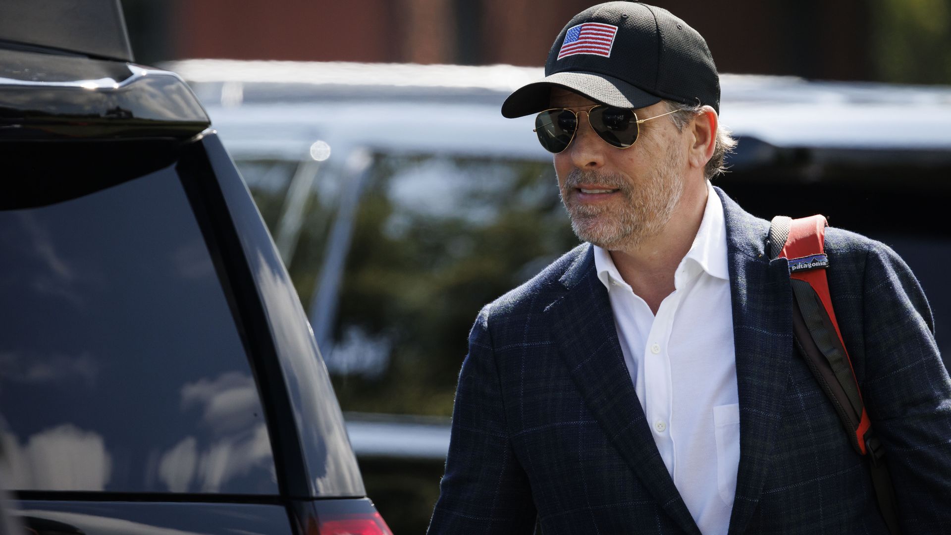 Hunter Biden, wearing a blue suit, and a black hat with an American flag on the front walks behind two SUVs.