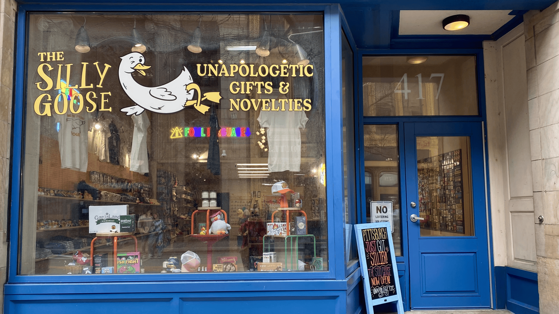 Blue storefront window for "The Silly Goose" gift shop with a white goose logo and gold text "UNAPOLOGETIC GIFTS & NOVELTIES"; interior shelves show shirts and toys; door reads 417.