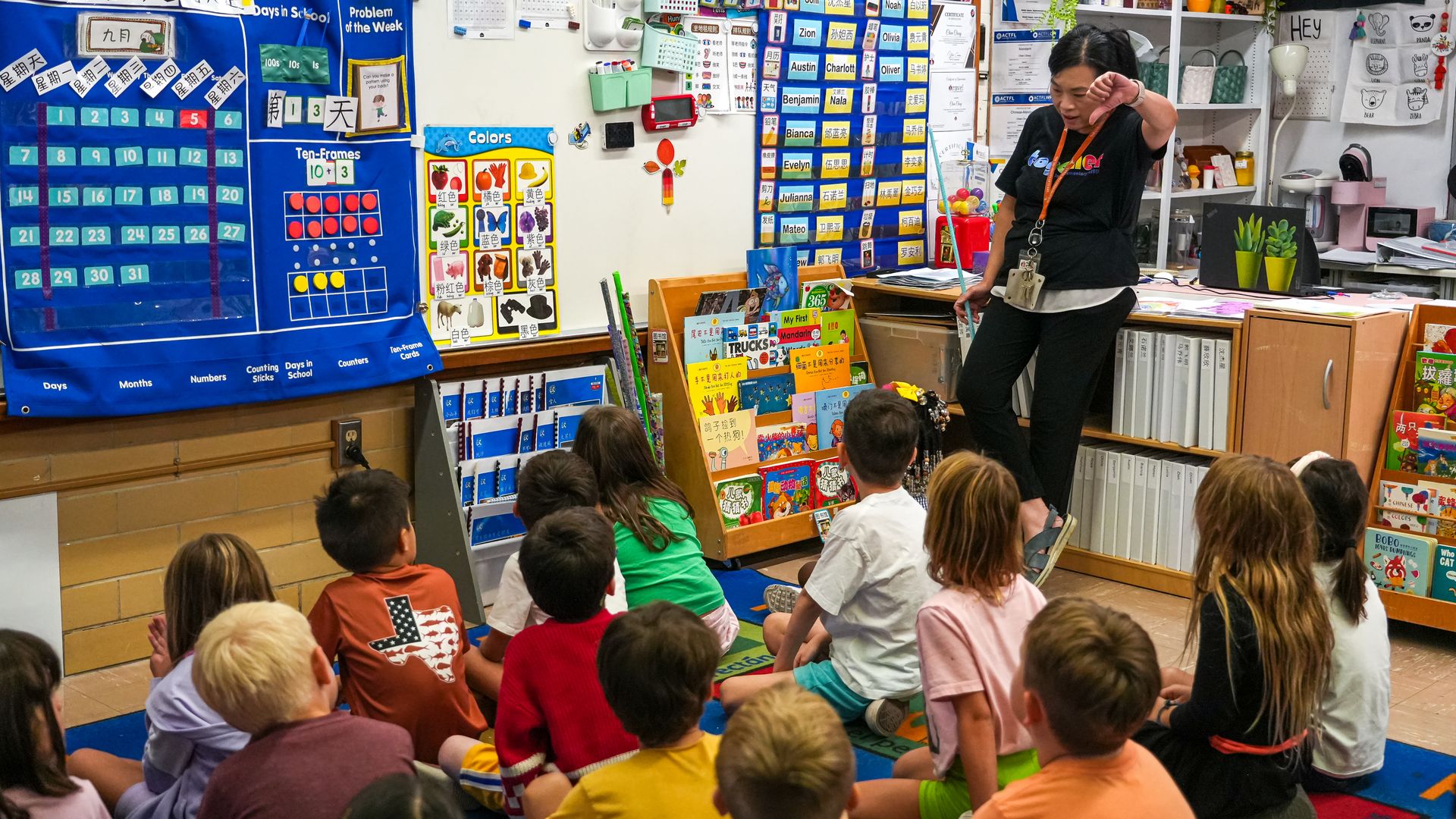 A teacher in a black shirt with an orange lanyard stands at the front of a bright classroom, gesturing toward seated children on a colorful rug, with blue charts and shelves of books along the walls.