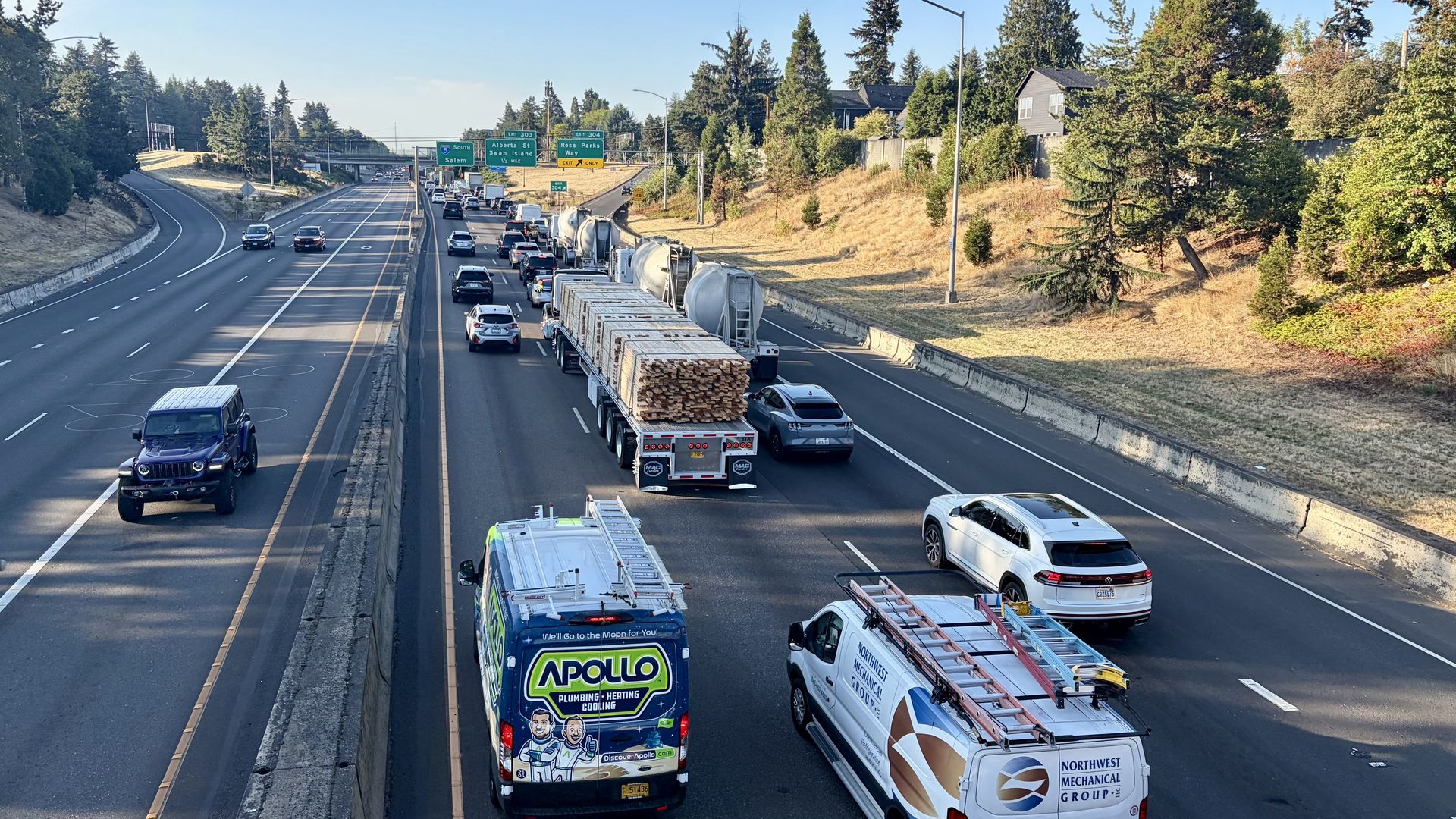 Aerial view of a busy multi-lane highway with cars and trucks, including a log truck carrying lumber and a tanker; green exit signs overhead; wooded hills and a few houses on the right.