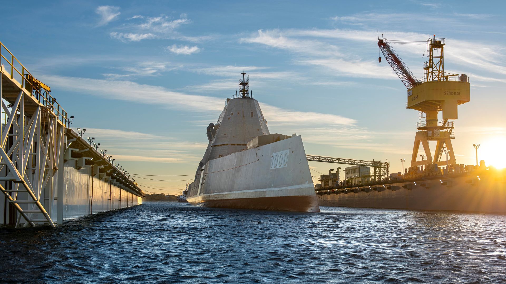 The USS Zumwalt sits in the water. Piers and other infrastructure are seen to the left and right of it. A crane is to the right and blocks a setting sun.