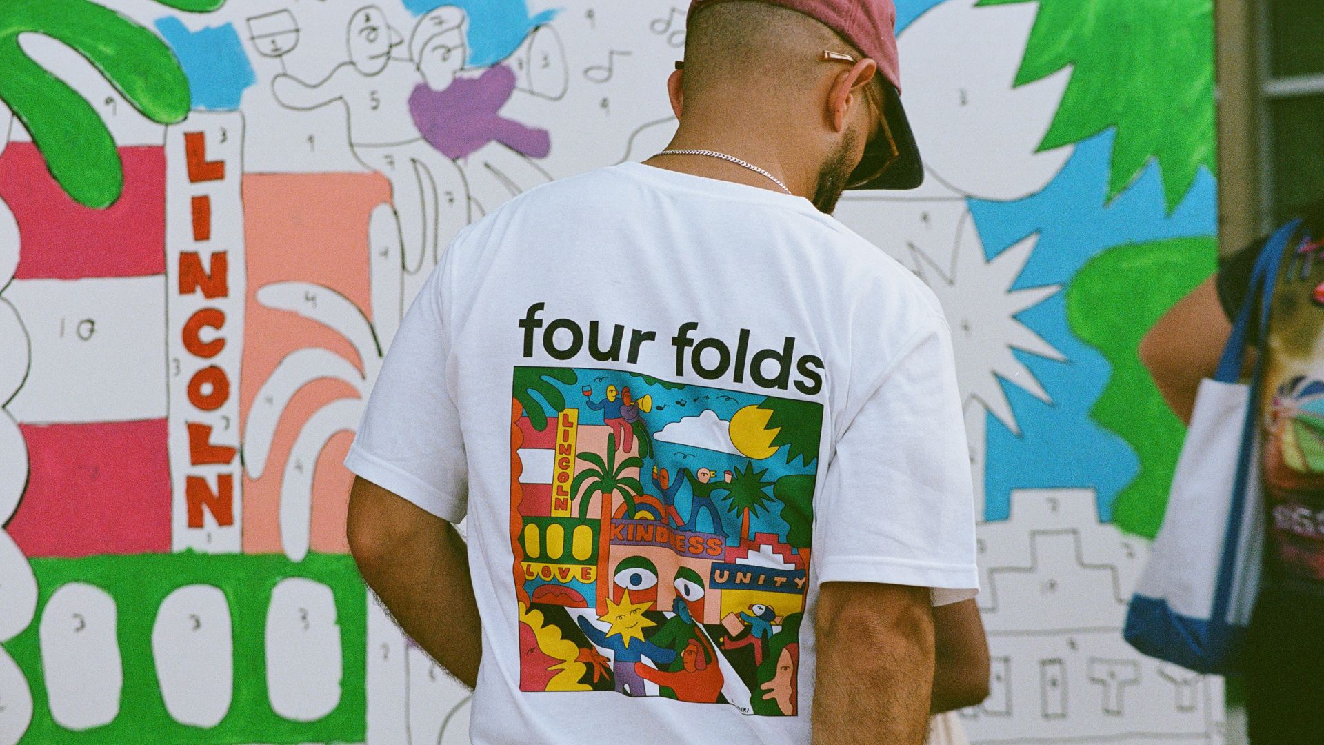 Back view of a man in a white T-shirt with a colorful graphic and the text "four folds". Bright graffiti wall in green and blue behind him.