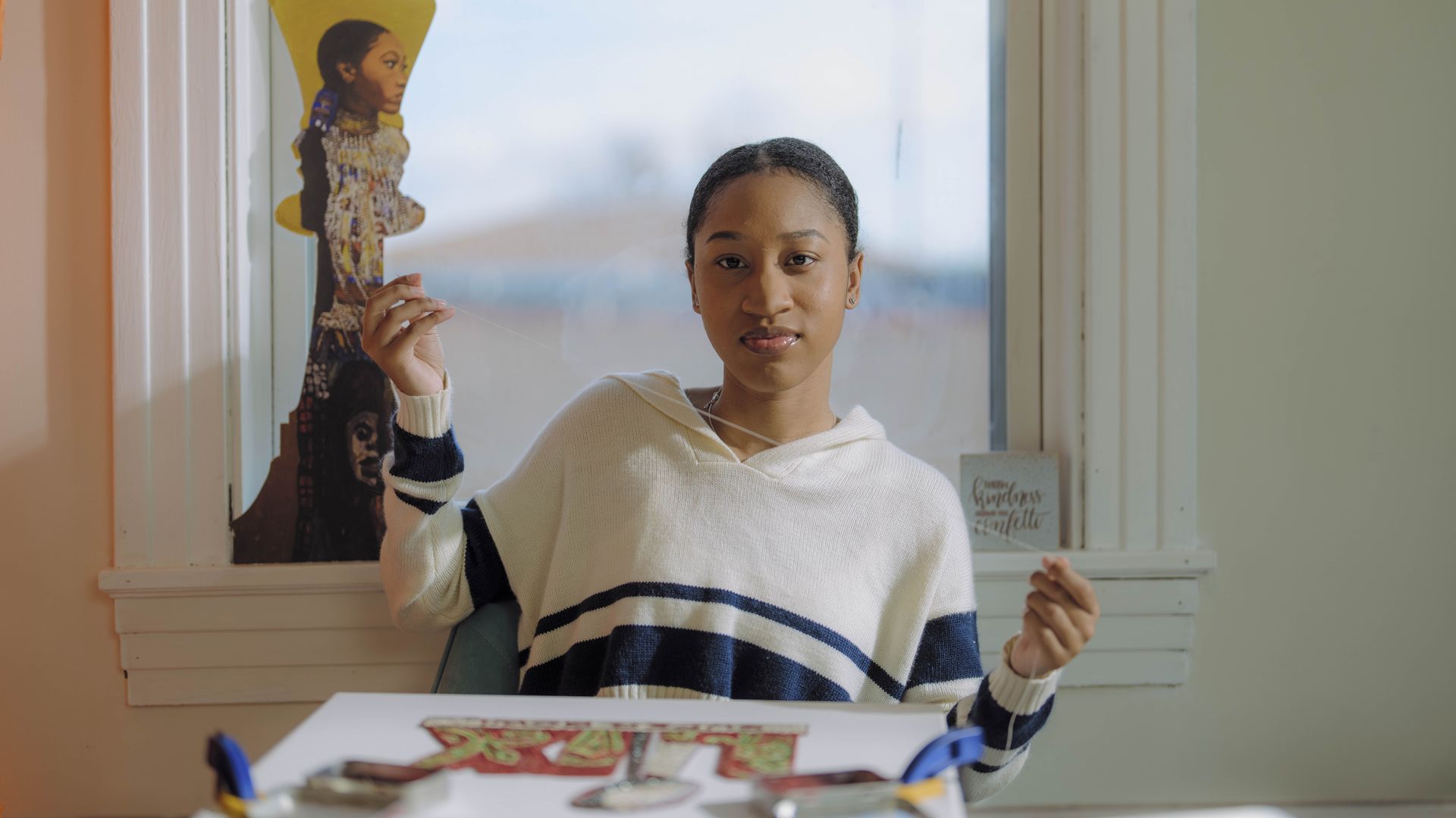 An image of Tahj Williams paused while sewing the Super Bowl LIX logo. She sits in front of a window and looks directly at the camera.